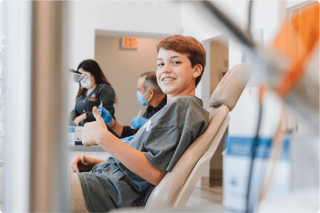 A young boy with orthodontic braces is sitting in a dental chair giving a thumbs up