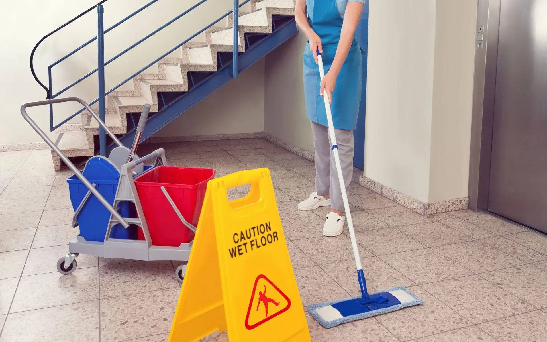A person mops a tiled floor, next to a wet floor sign and cleaning cart.