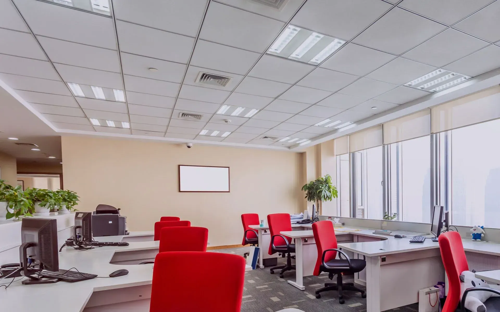 An office space with red chairs, beige walls, and white desks under a grid ceiling with bright lights.