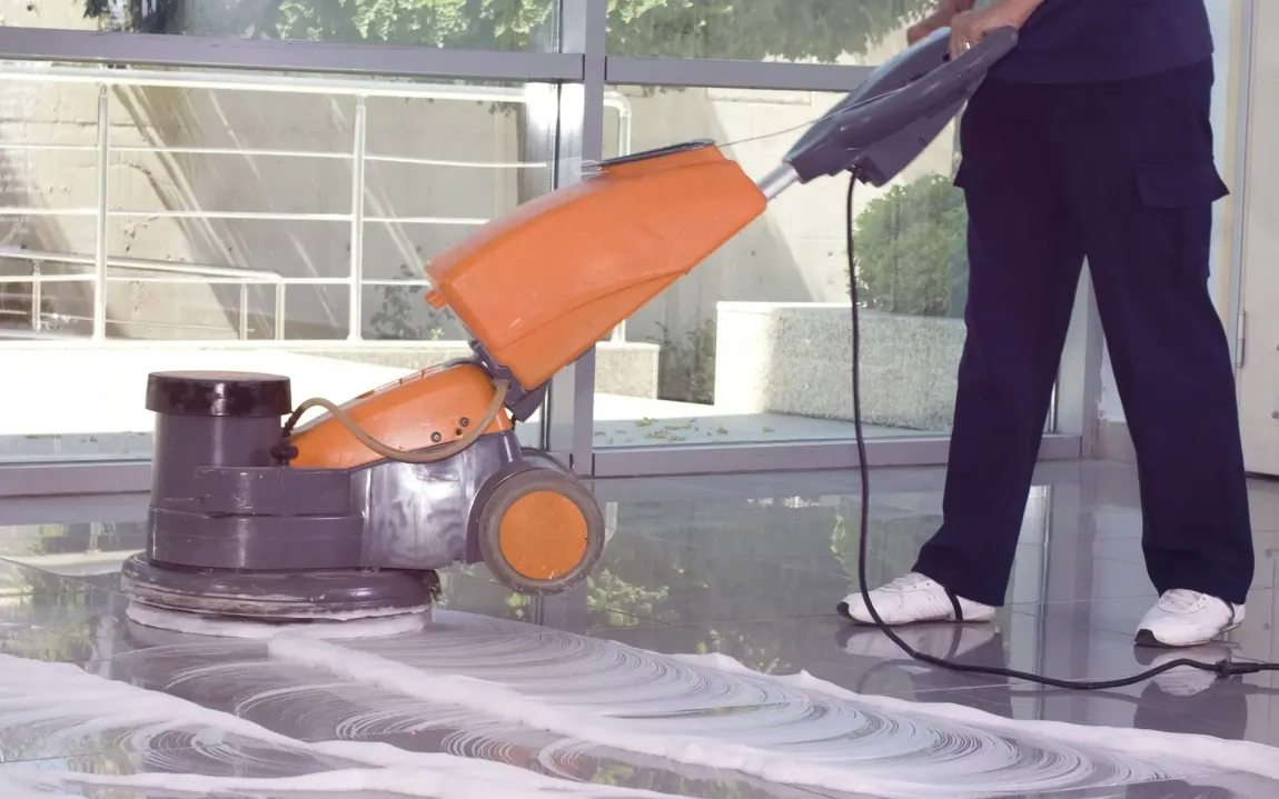 Person cleaning floor with an orange and gray floor buffer in an office setting.