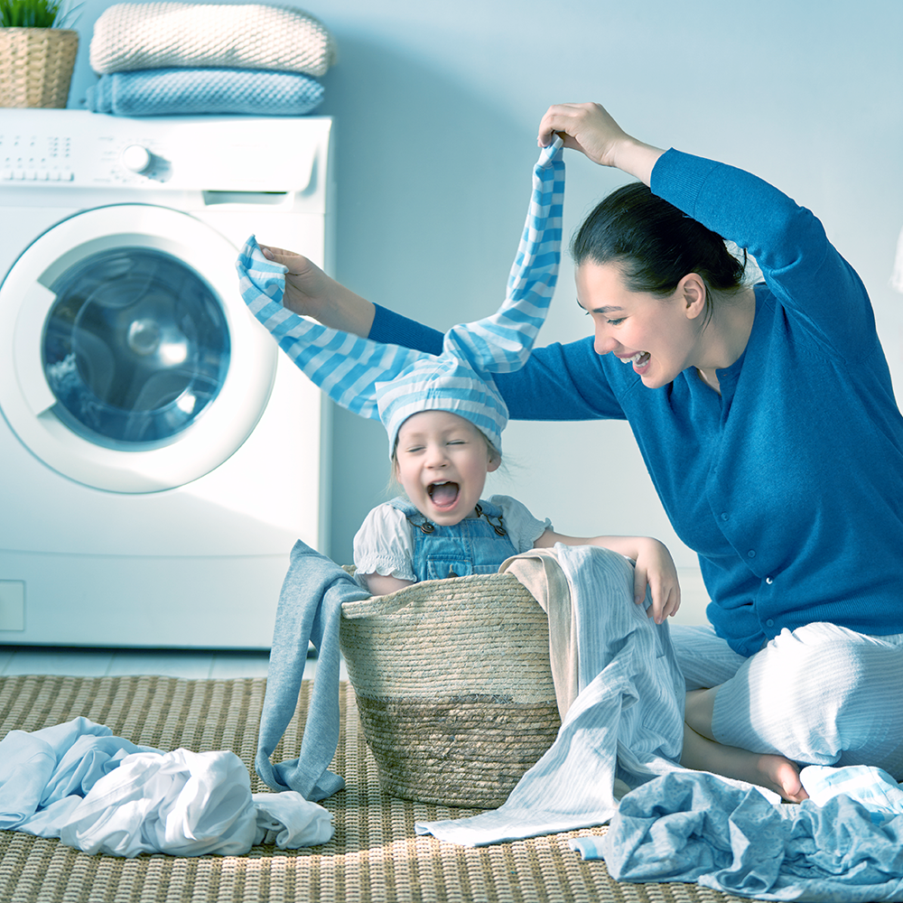 אישה משחקת עם ילד, חובשת כובע מפוספס כחול על ראשו, ליד סל כביסה ומכונת כביסה.