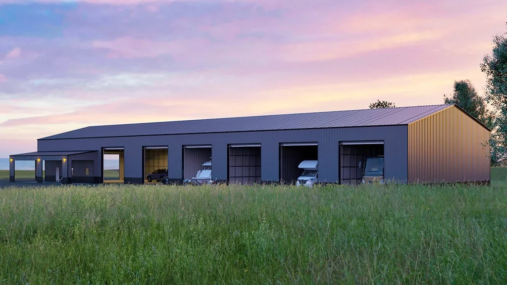 A modern dark gray metal building with open bays, housing vehicles, in a field at dusk.
