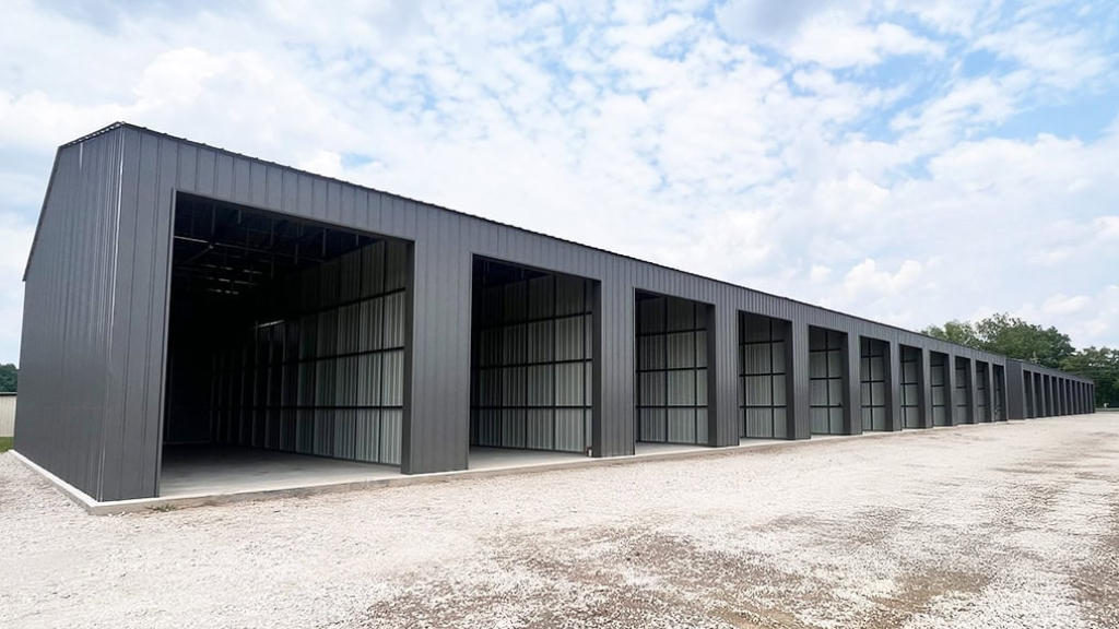 Tan and red metal building with three garage door openings, red roof, and concrete slab.