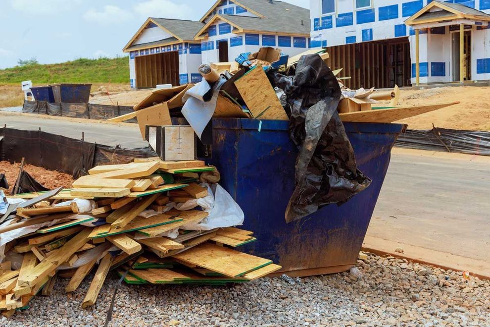 Blue dumpster overflowing with construction debris, in front of partially built houses.