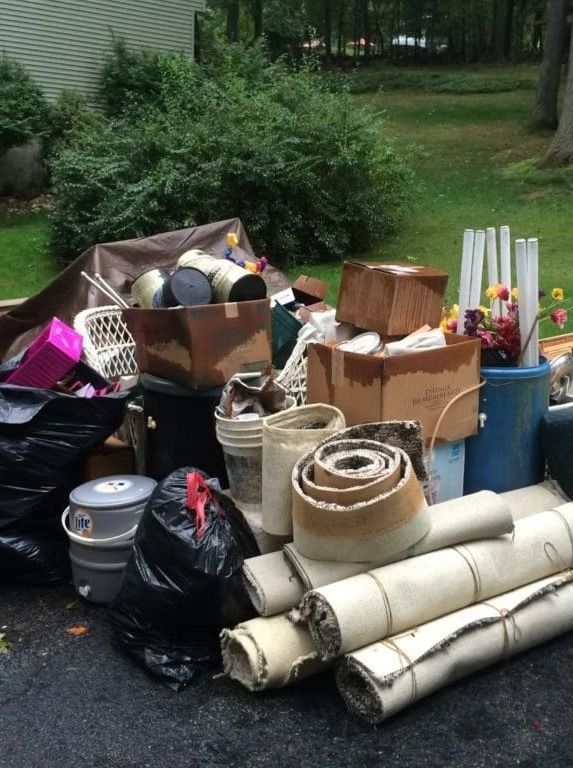 Pile of discarded items, including boxes, rolls of carpet, and trash bags, in a yard.