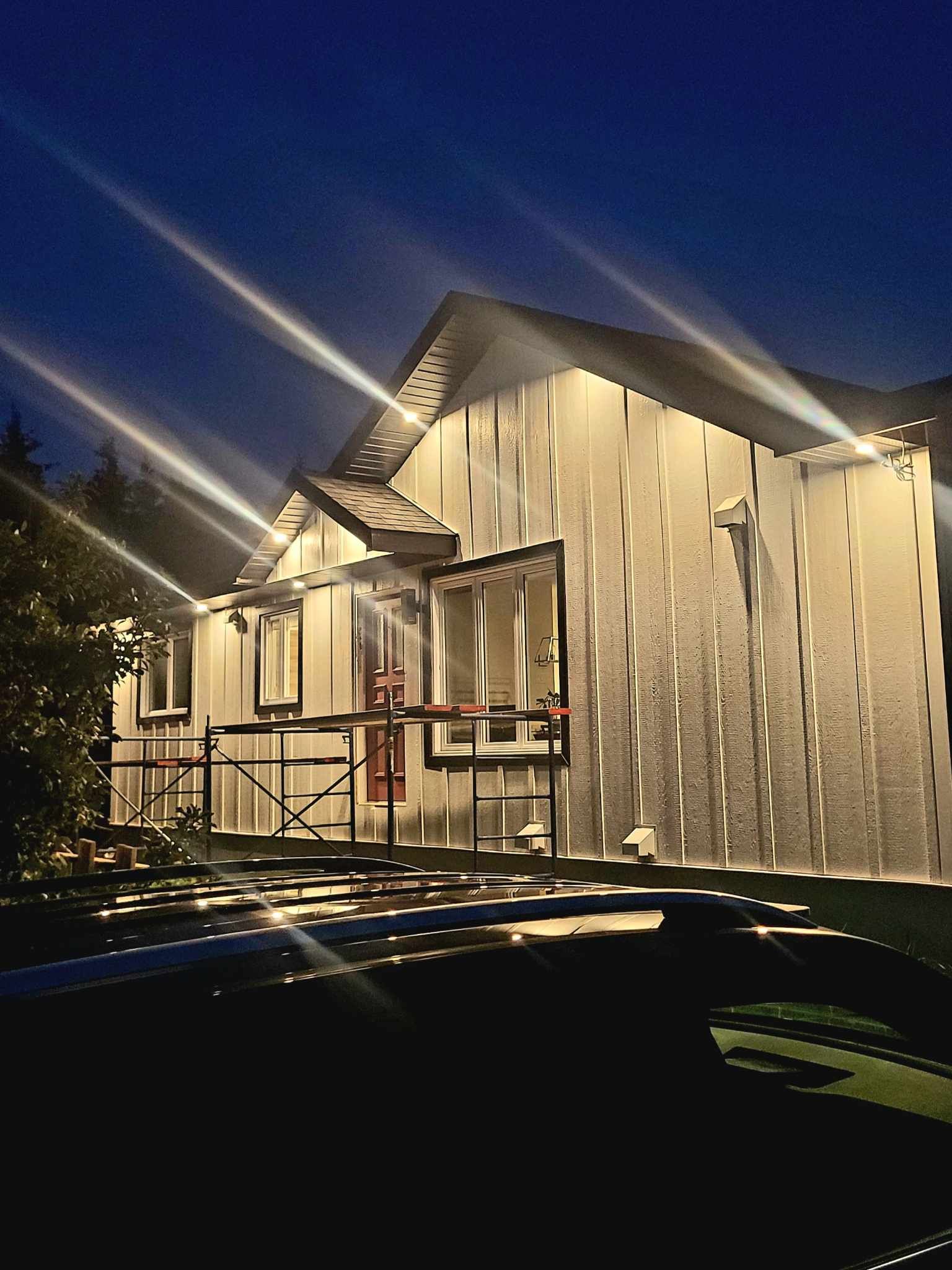 Façade de la maison la nuit, illuminée par des lumières vives. Ciel bleu foncé, pelouse devant.