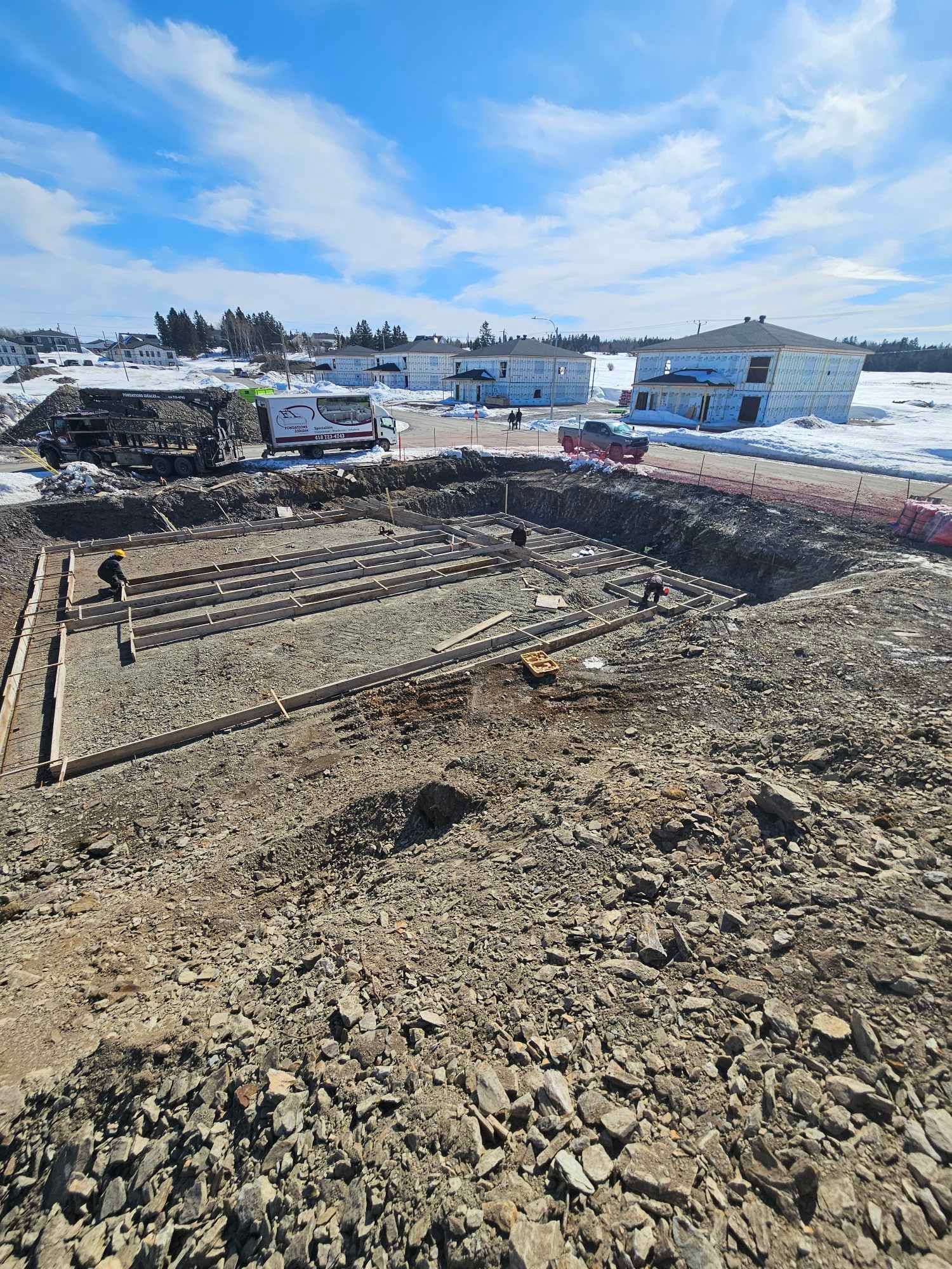 Chantier de construction avec fondations excavées, bois de construction et neige éparse.