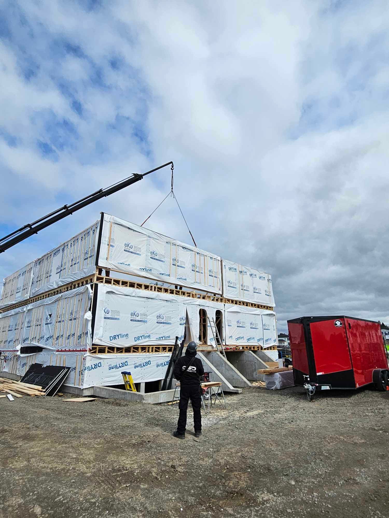 Chantier : Grue soulevant un module de construction. Un homme en noir se tient près d’une structure à deux étages