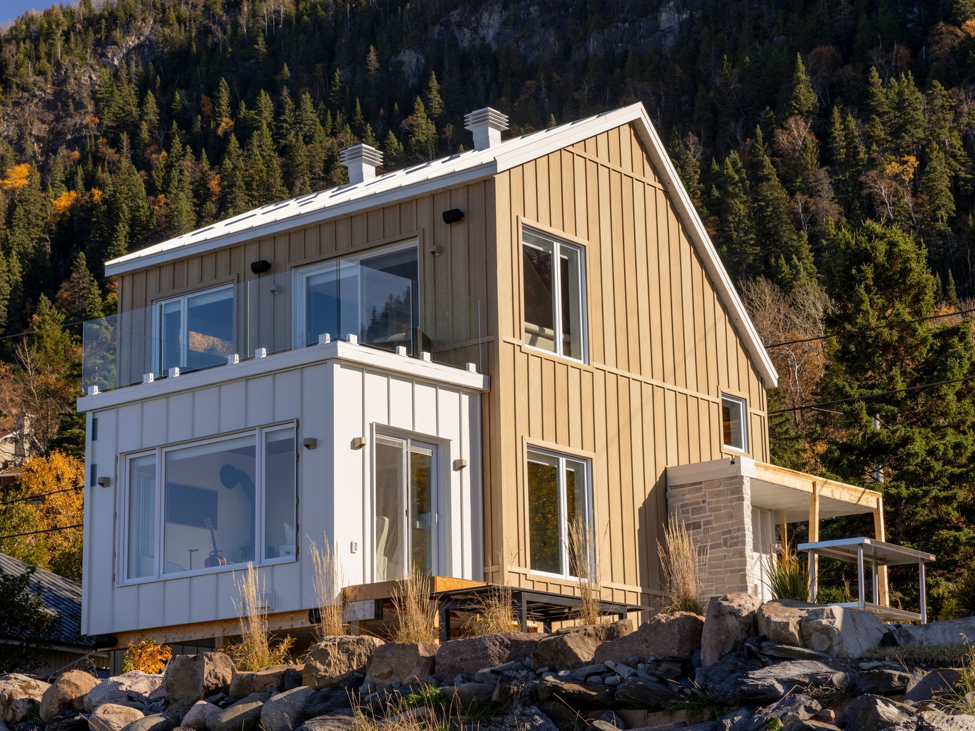 Maison moderne à deux étages avec un revêtement extérieur brun clair et blanc, des balcons vitrés et un premier plan rocheux