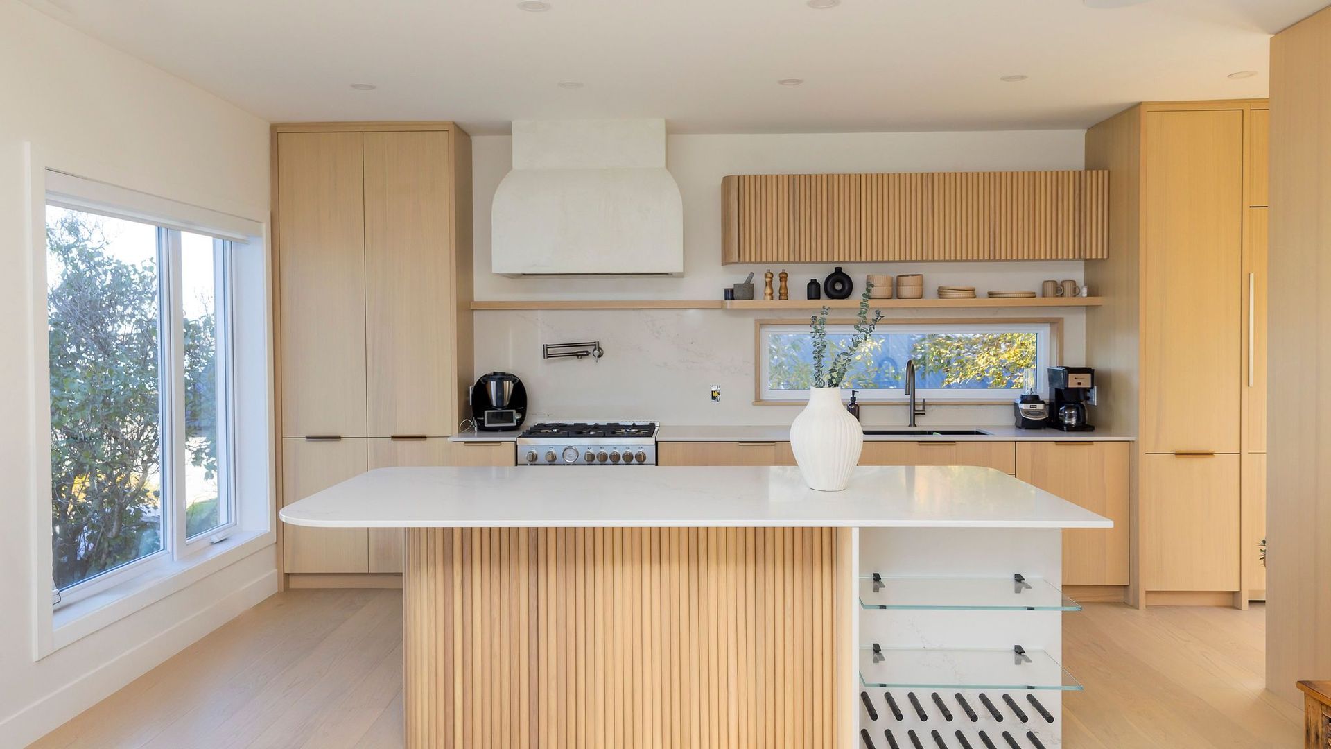 Cuisine moderne avec armoires en bois clair, comptoirs blancs et îlot avec panneaux de bois cannelés.