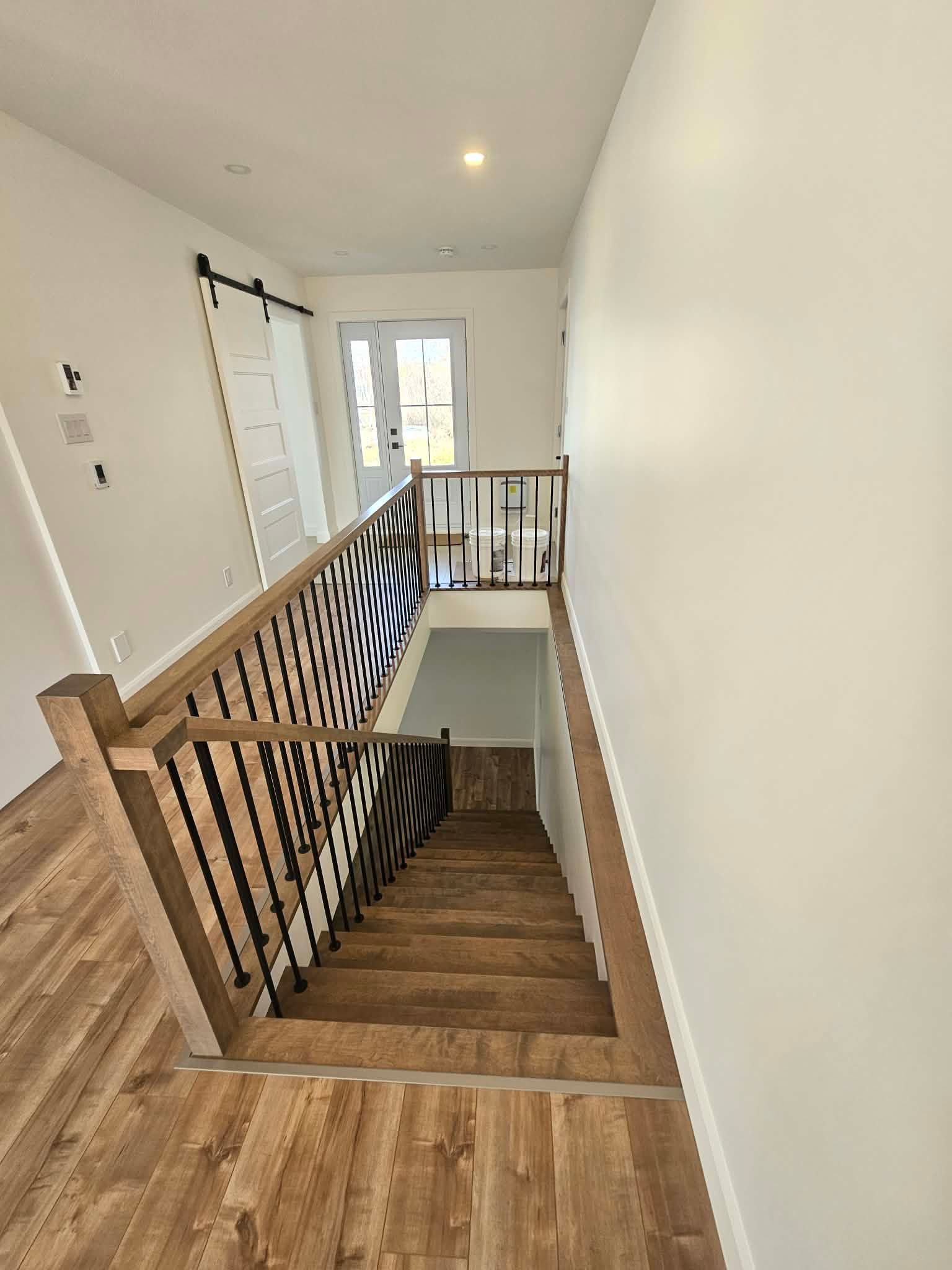 Un escalier en bois avec des rampes en métal noir descend d'un couloir aux murs blancs et au sol en bois.