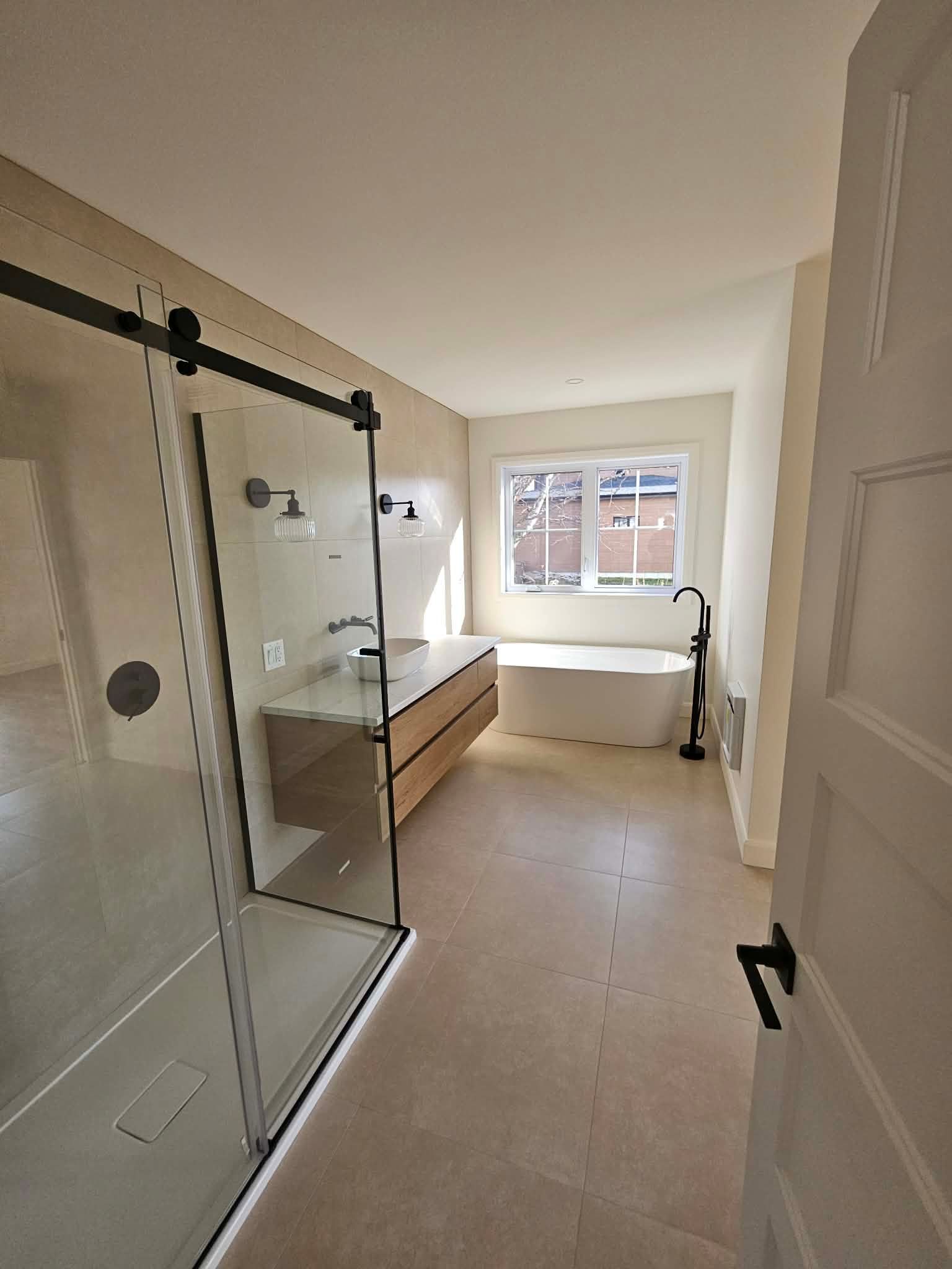 Salle de bains moderne avec douche en verre, meuble-lavabo en bois clair, baignoire et fenêtre.