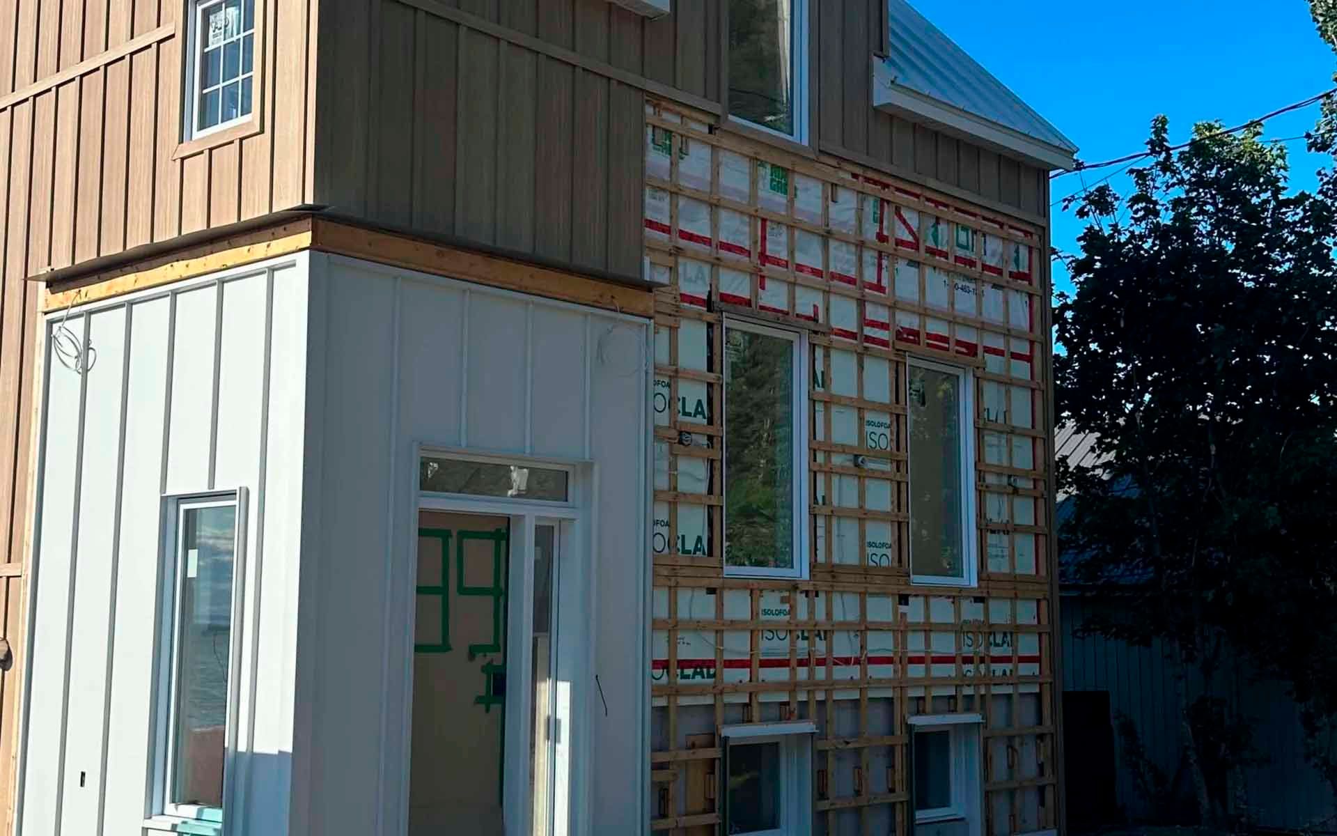 Une maison à deux étages en construction, dont le revêtement extérieur est partiellement installé ; le ciel est bleu.