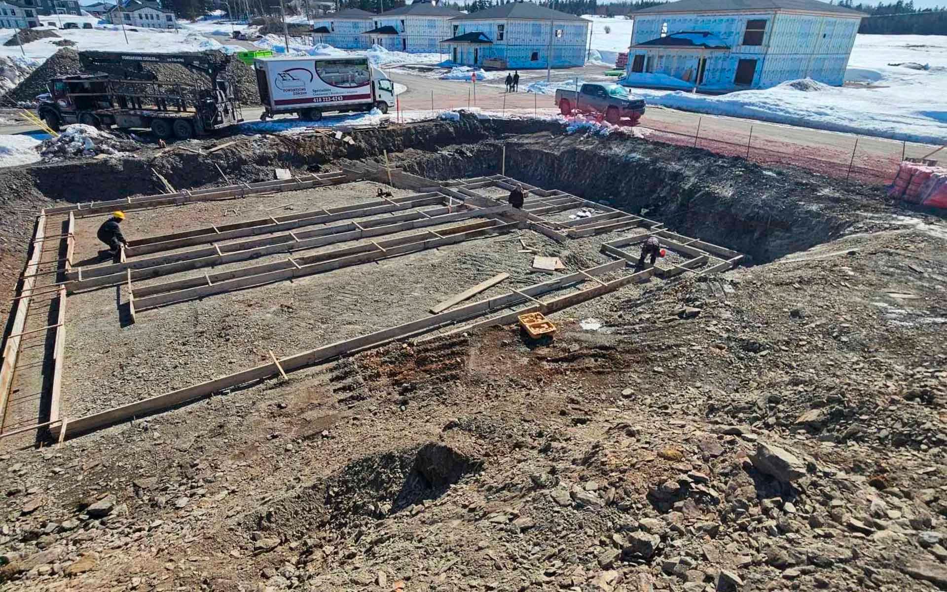 Chantier de construction avec des ouvriers dans une fosse de fondation, des coffrages en bois et une base de gravier.