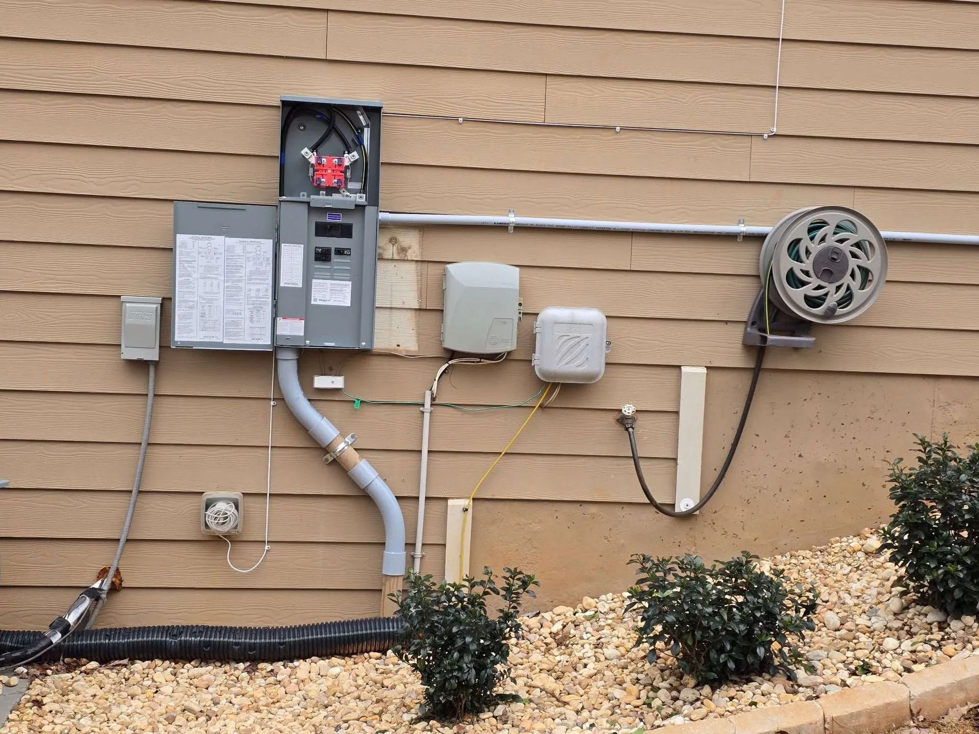 Electrical panel, conduit, and related components mounted on a tan siding exterior wall.