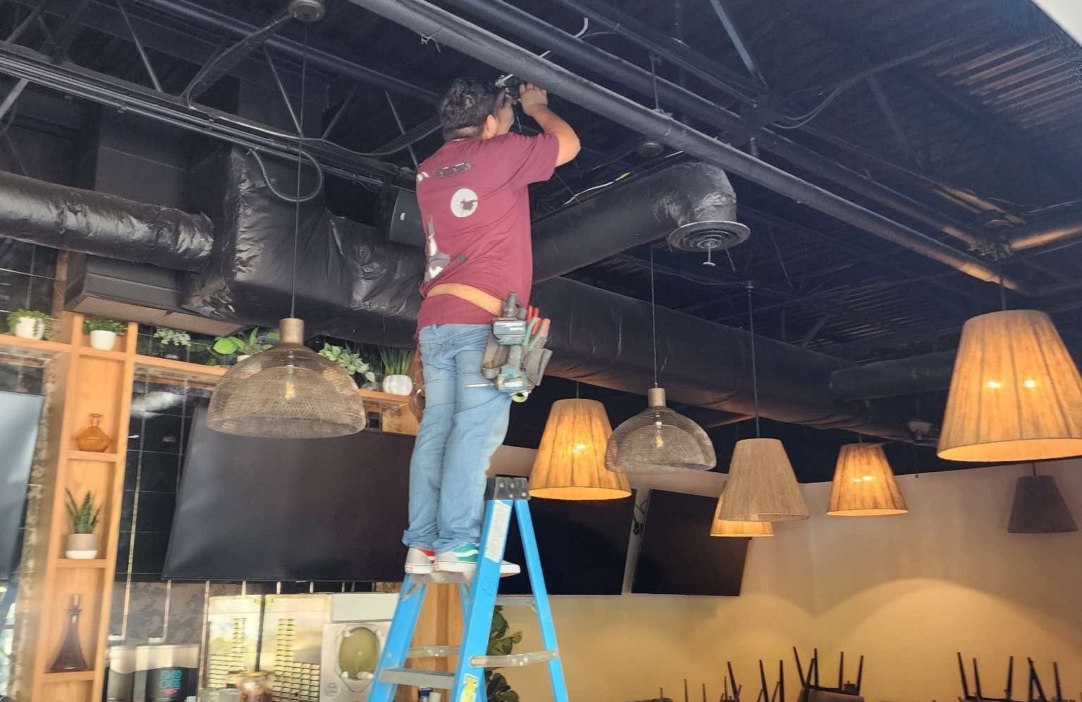 Man on a ladder installing something on a black ceiling in a restaurant, with hanging lights and ductwork visible.