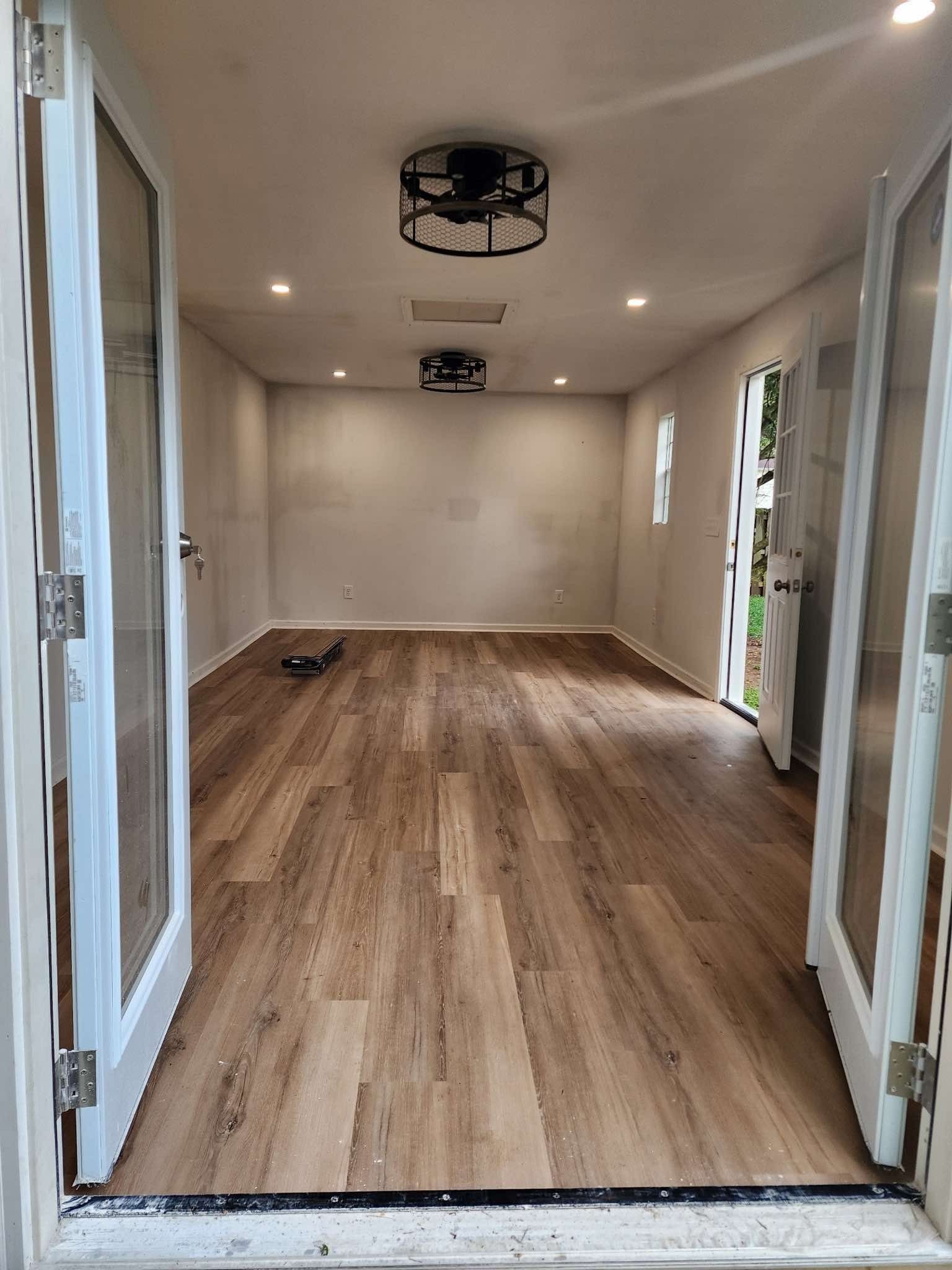 Empty room with wooden floors, white walls, and glass doors. Ceiling light fixtures are visible.