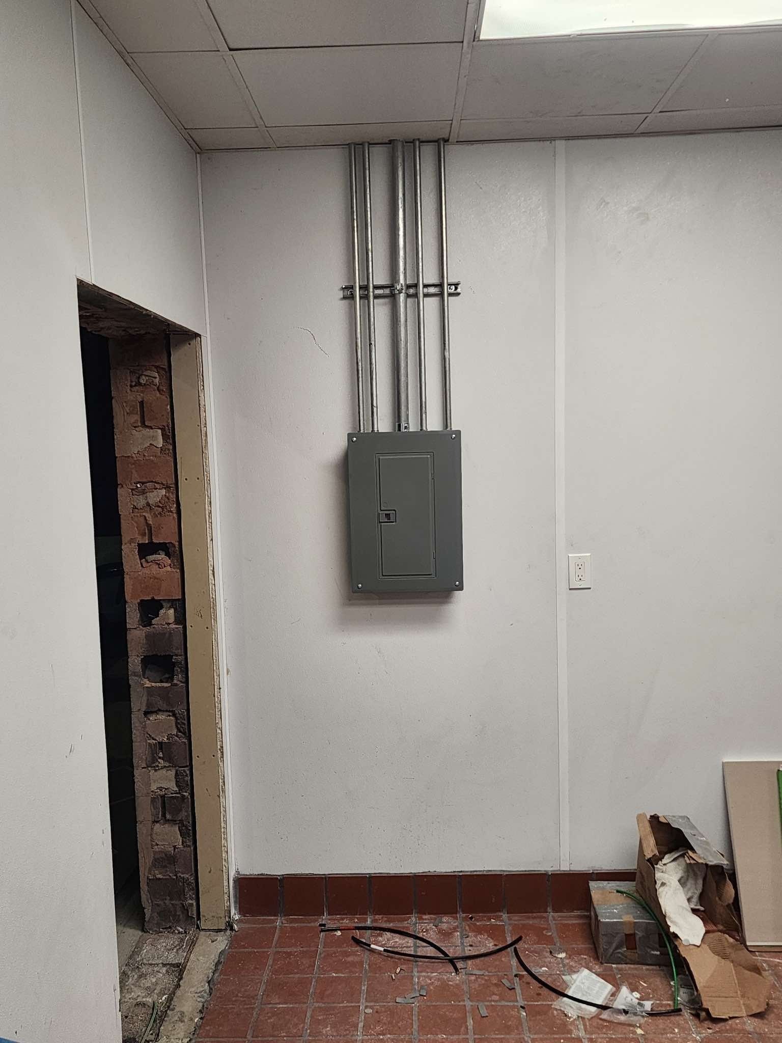 Electrical panel on a white wall with metal conduit. Door frame on the left, debris on the floor.