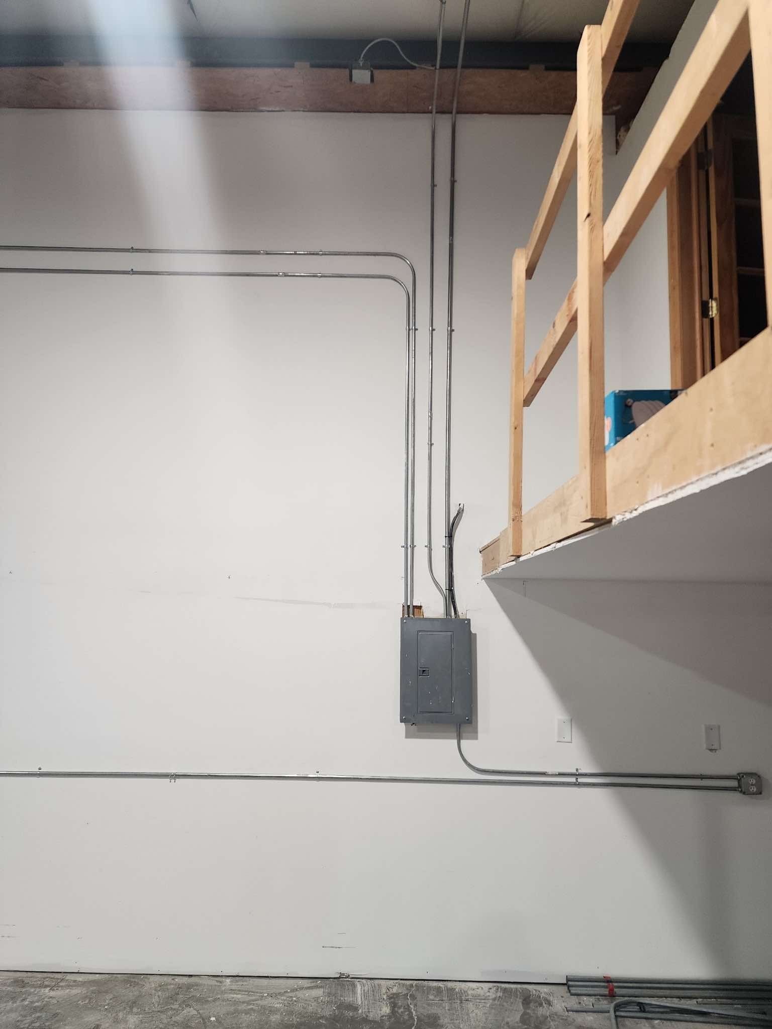 Electrical conduit and panel on a white wall below a wooden platform.