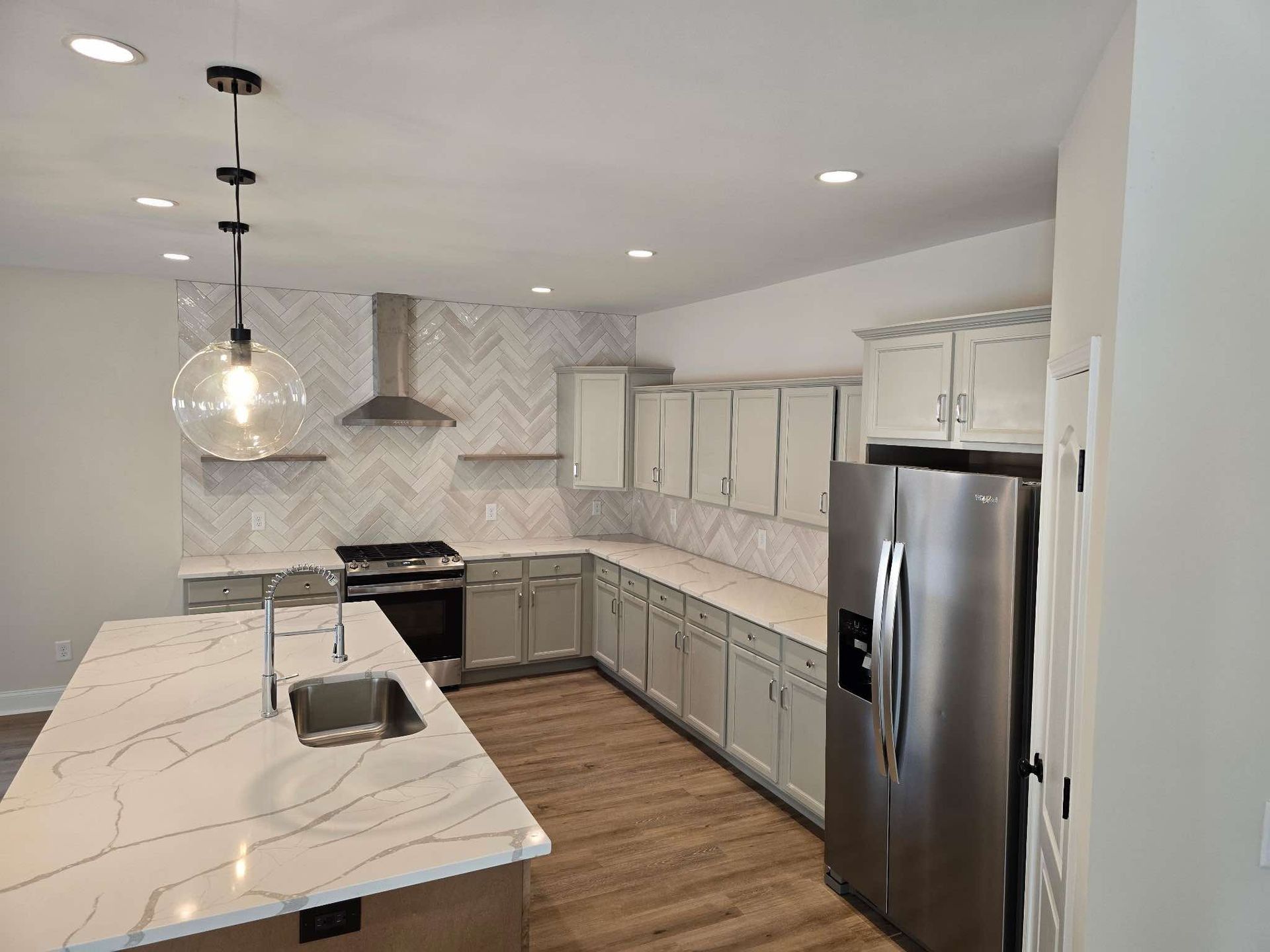 Modern kitchen with gray cabinets, marble countertops, stainless steel appliances, and a herringbone backsplash.