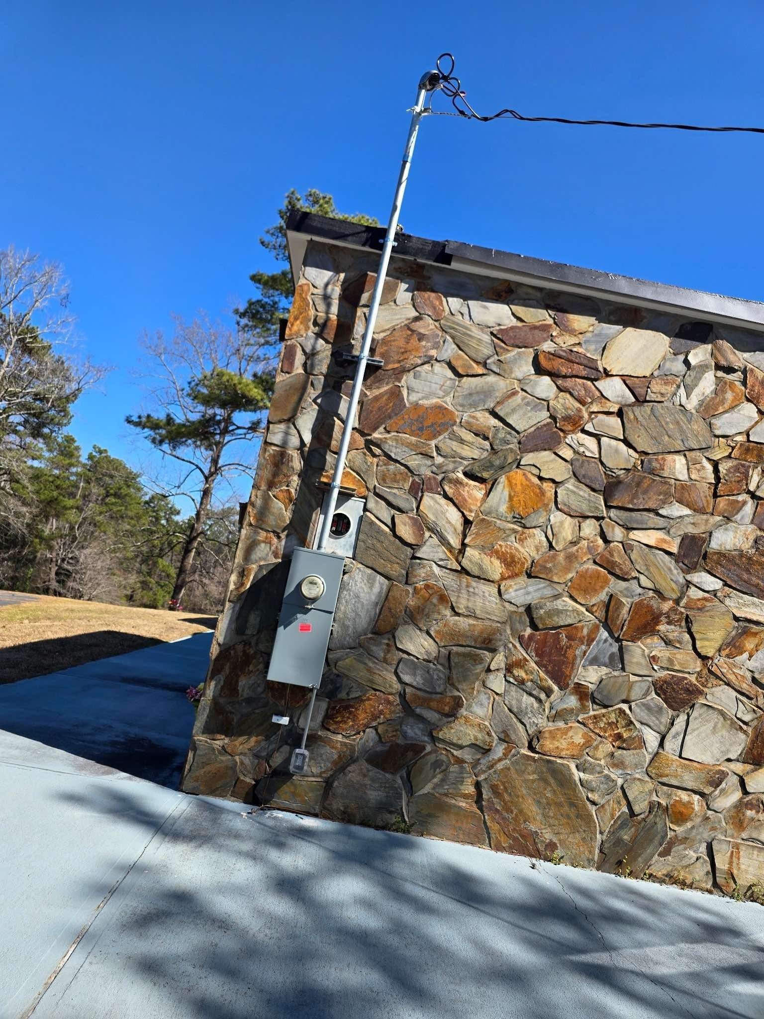 Electrical meter box and conduit mounted on a stone wall. Power lines visible against a blue sky.