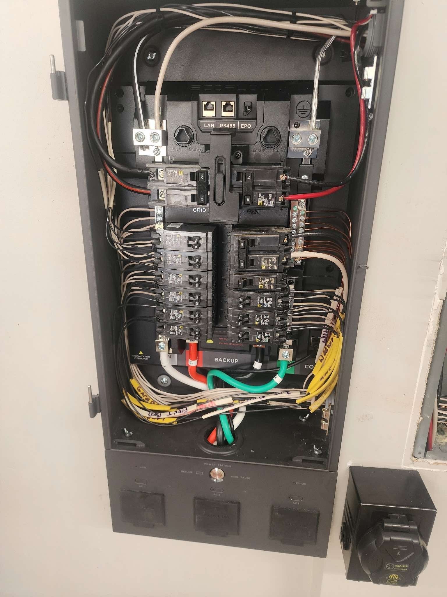 Electrical panel with wires and circuit breakers, mounted on a wall.