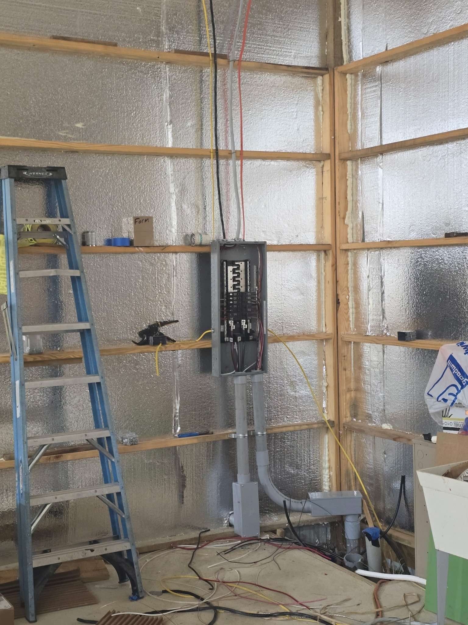 Electrical panel installation in a wood-framed room with ladder, wiring, and insulation visible.