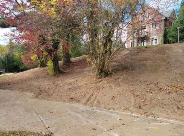 A house is sitting on top of a hill next to a sidewalk and trees.