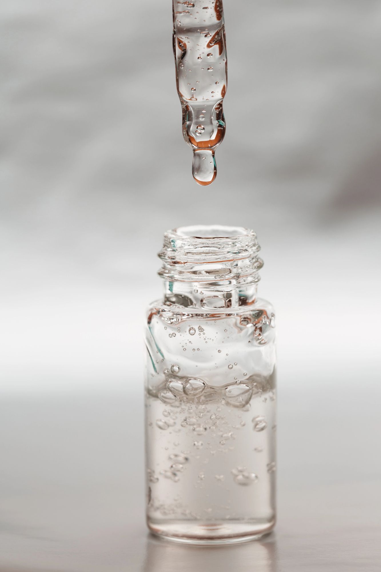 Gotero dispensando líquido transparente en una pequeña botella de vidrio, burbujas en el interior, fondo gris.