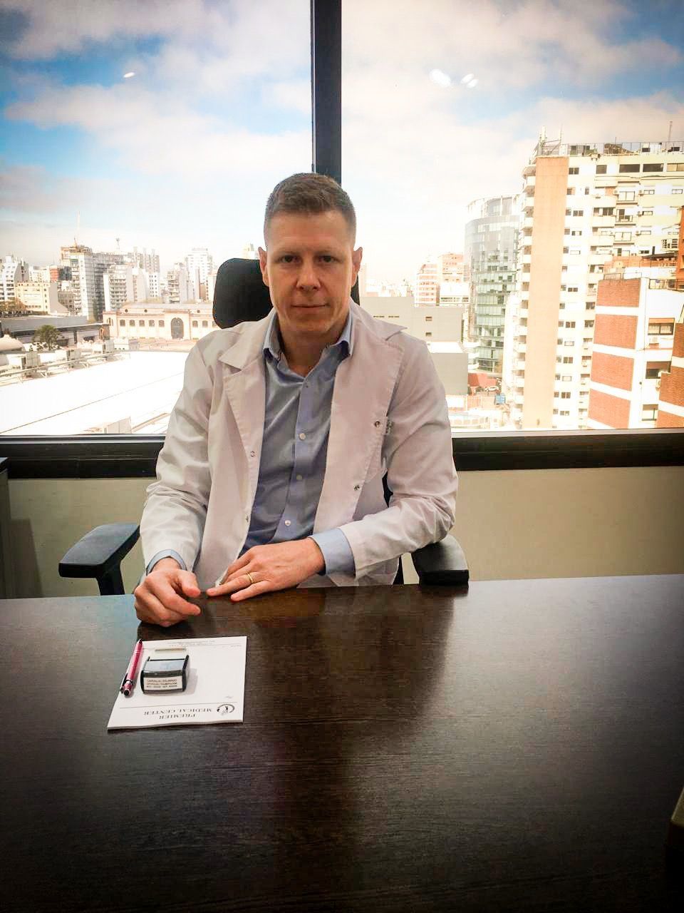 Un médico examina el tobillo de un paciente. La pierna descansa sobre una toalla blanca. El médico lleva una bata blanca.