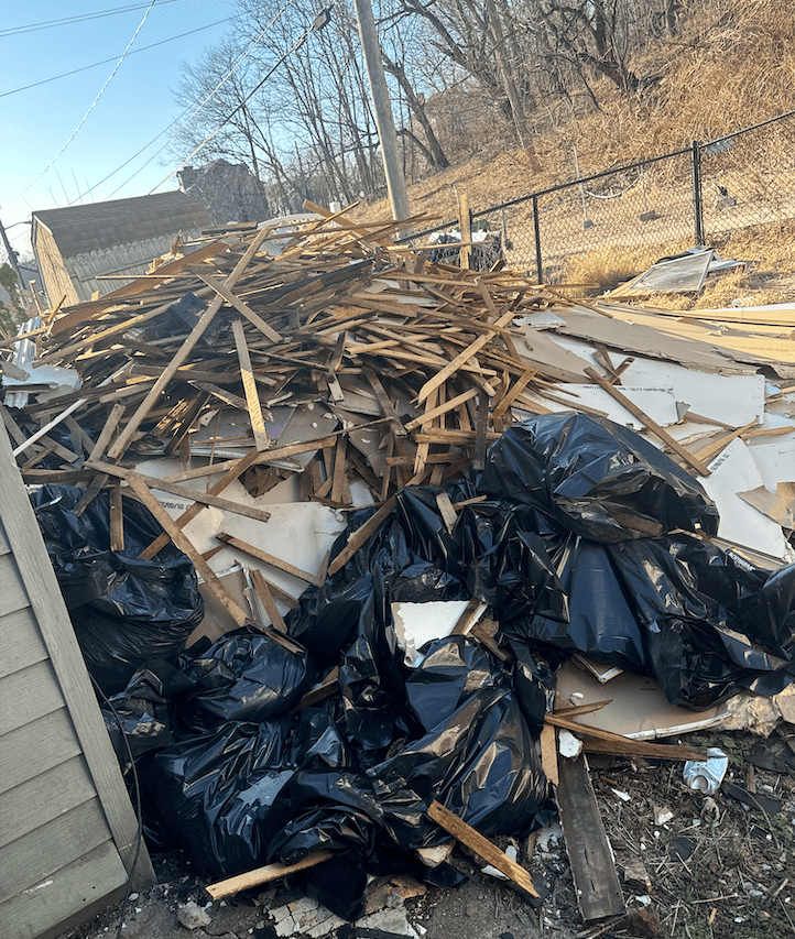 Pile of construction debris, wood scraps, and black trash bags in an outdoor setting.