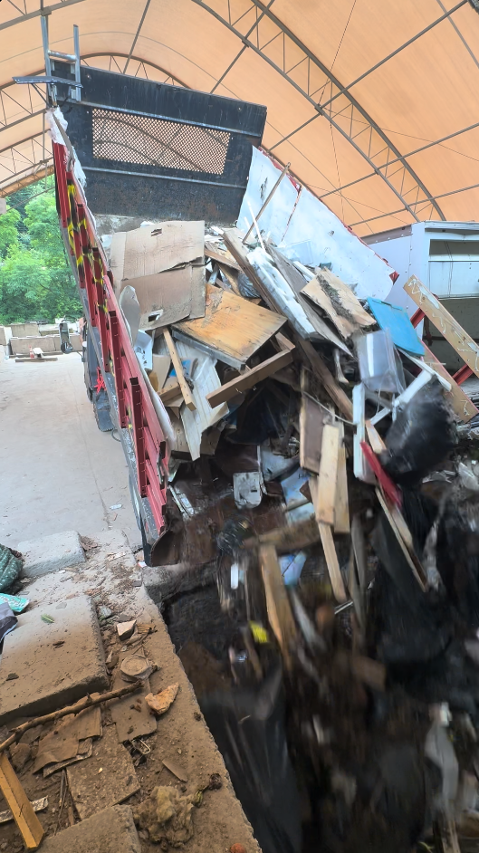 Truck bed filled with debris, tilting to unload. Wood, plastic, and trash are visible. Under a warehouse roof.