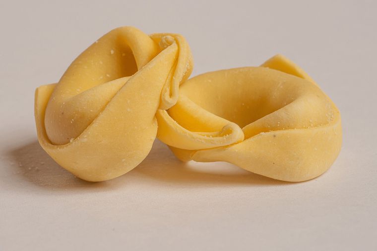 Due pezzi di pasta tortellini crudi, gialli, in primo piano su uno sfondo chiaro.