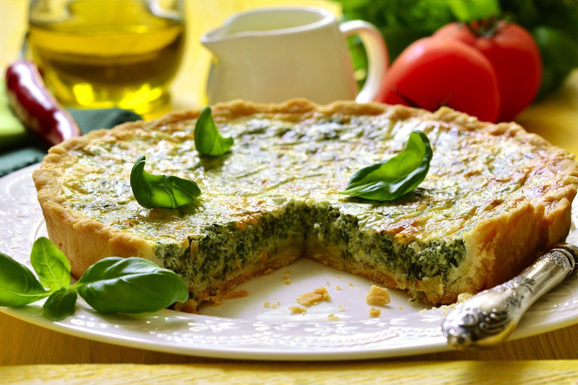Quiche di spinaci su un piatto bianco con una fetta tagliata, guarnita con foglie di basilico, vicino a pomodori e olio d'oliva.