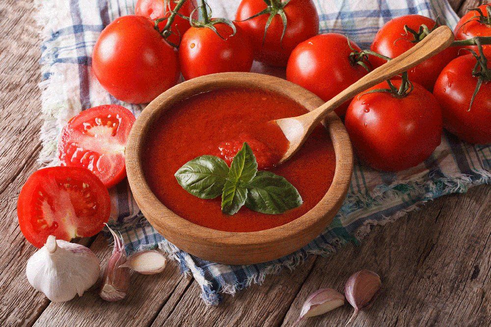 Zuppa di pomodoro in una ciotola di legno con pomodori, basilico, aglio e un cucchiaio su una tovaglia a quadretti.
