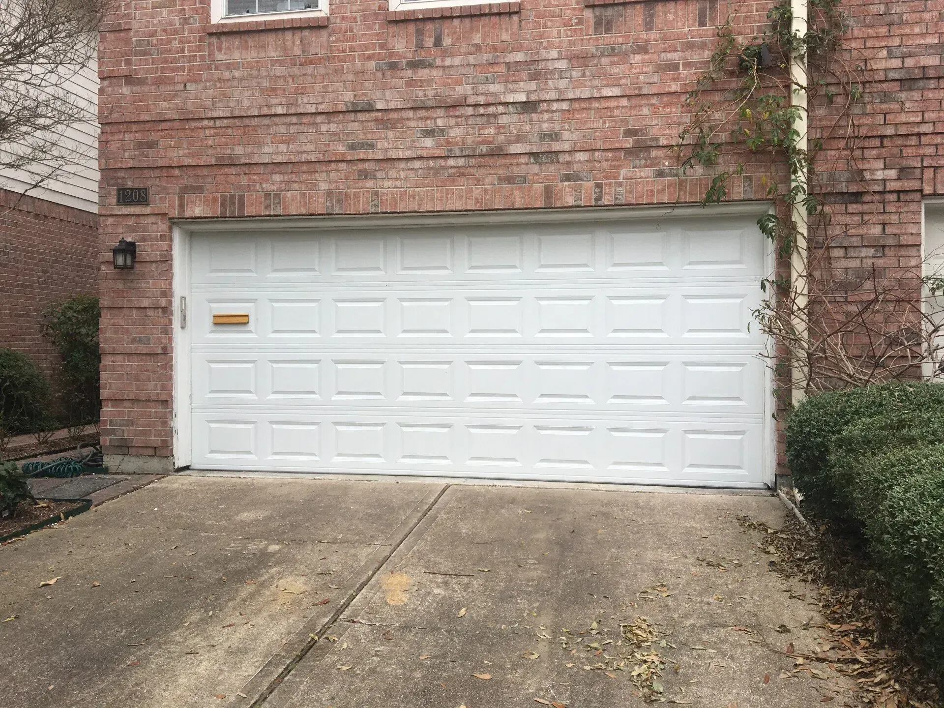 A white, rectangular paneled garage door on a brick house with a concrete driveway in front.