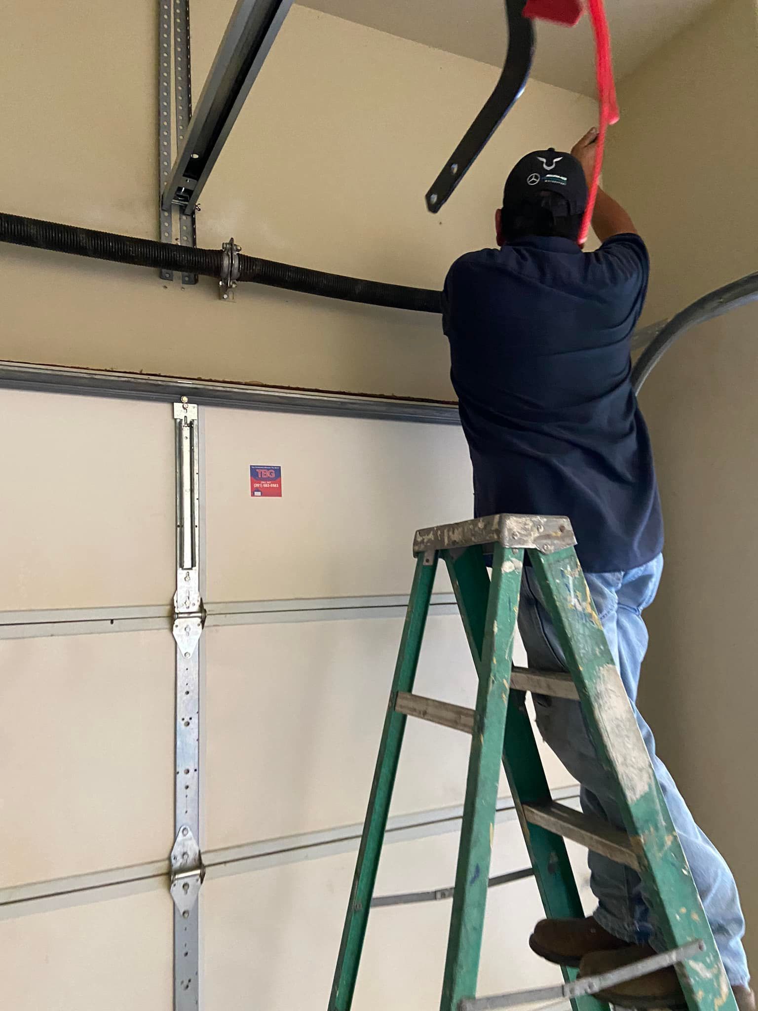 A person in a dark shirt on a ladder adjusting the release cord of a garage door opener.