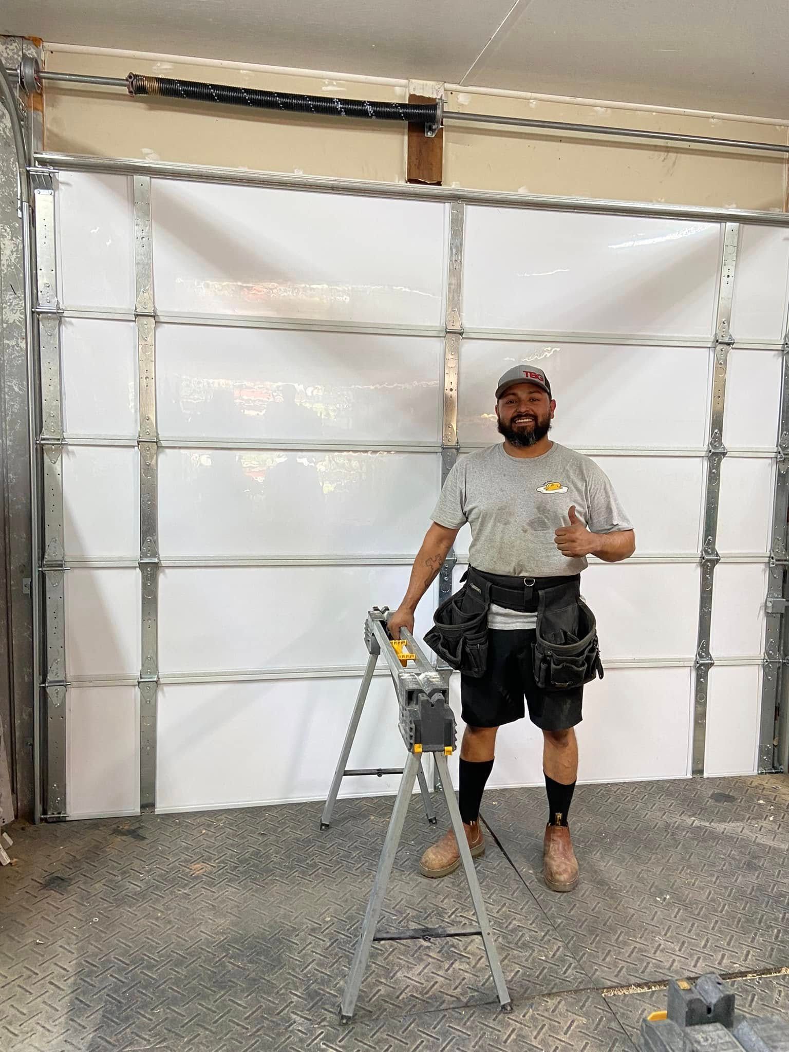 A smiling technician wearing a tool belt stands in a garage in front of a newly installed white metal garage door.