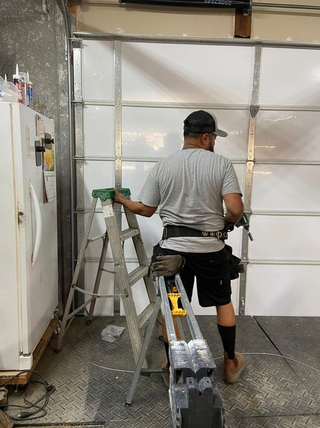 A worker in a gray t-shirt stands on a stepladder while installing or repairing a white garage door in a workshop.