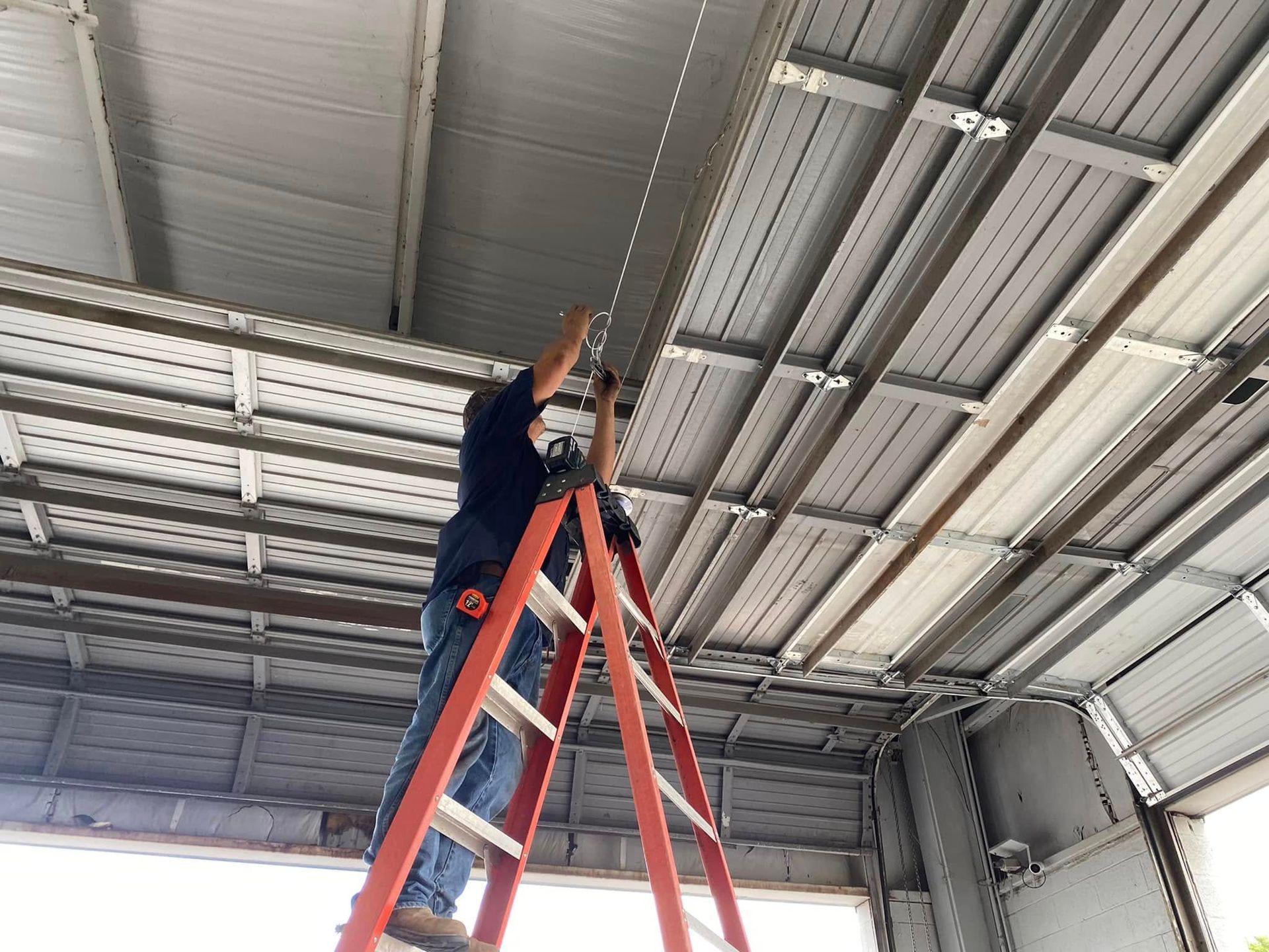 A person standing on an orange stepladder, working on the ceiling structure of an open, metal-framed garage.