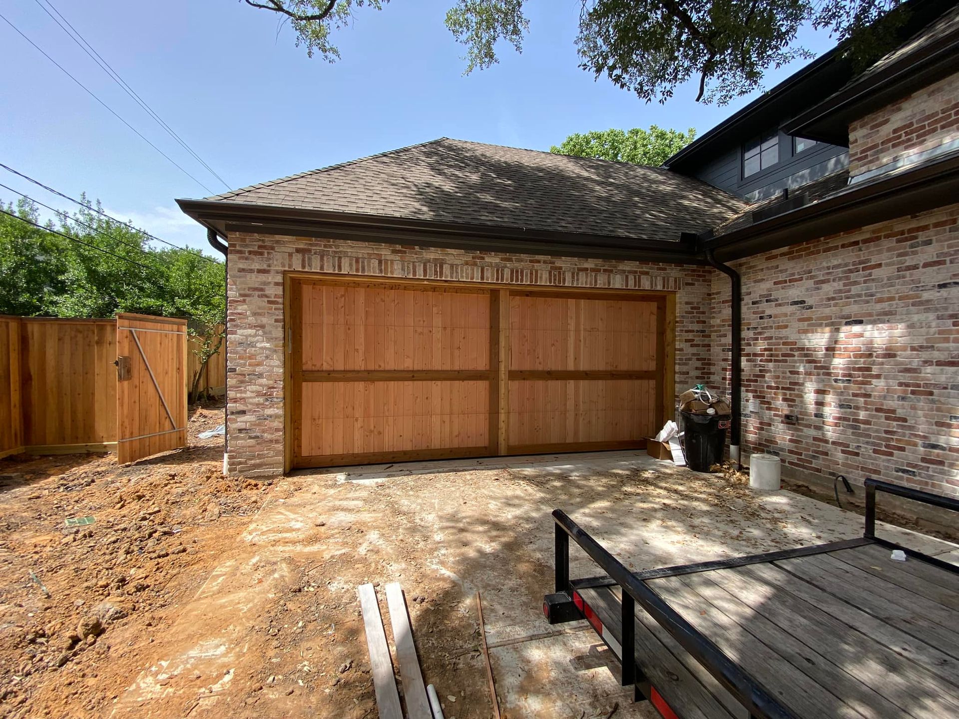 Exterior view of a brick house featuring a two-car wooden garage door, adjacent wooden fencing, and a utility trailer.