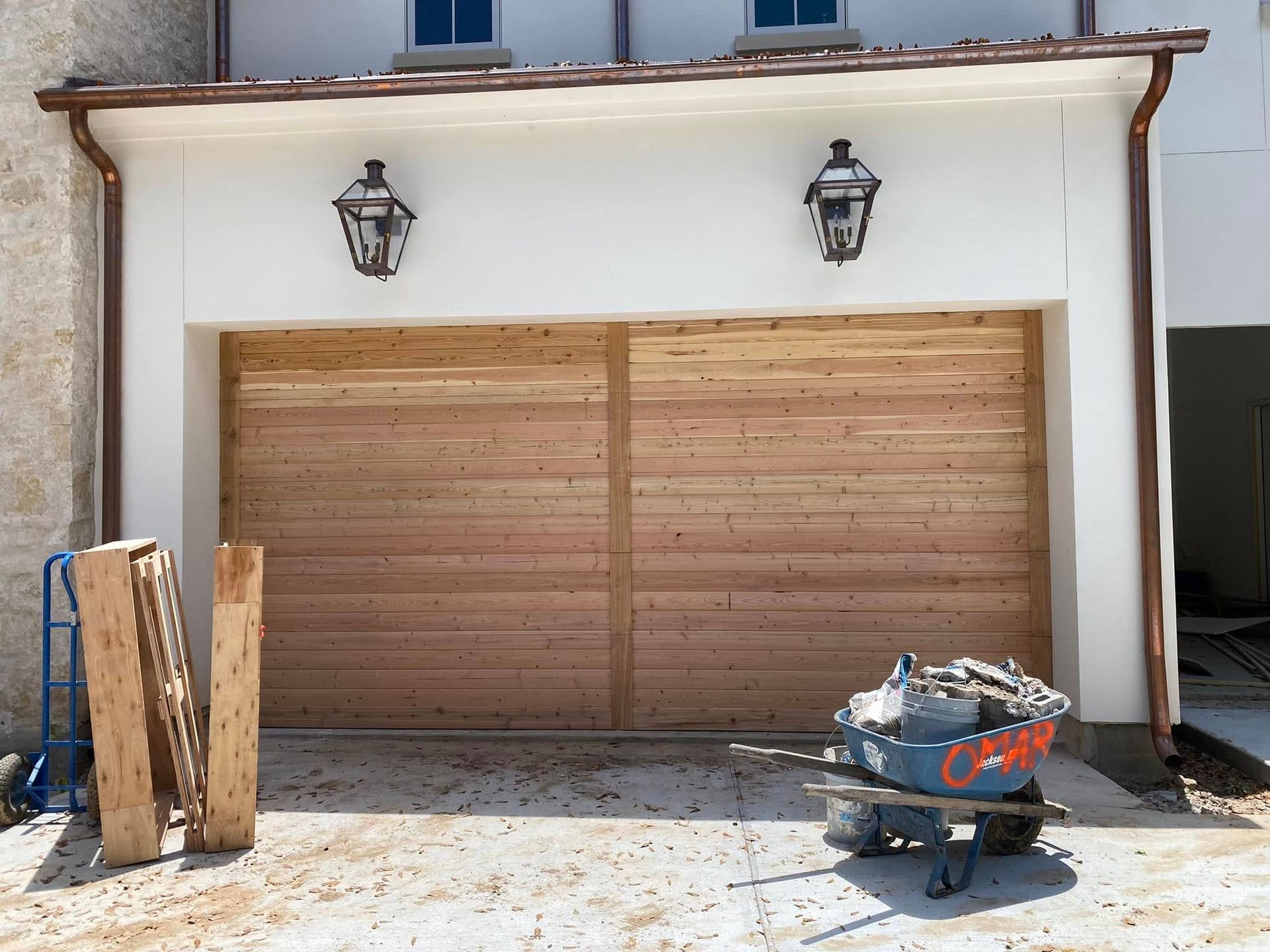 A modern wooden garage door set in a white exterior wall with two black wall lanterns and copper gutters.