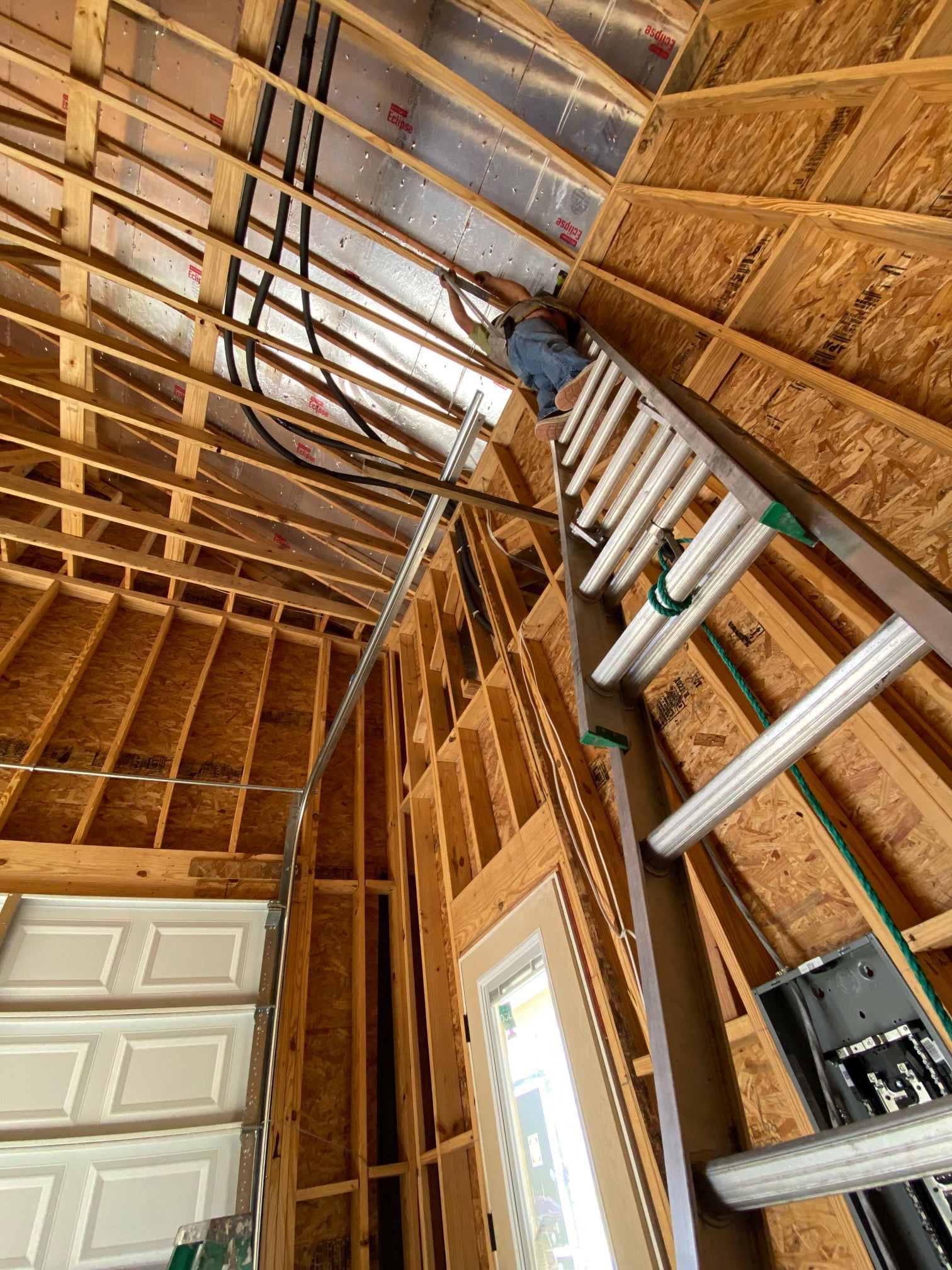 A person on a ladder installs wiring near the ceiling rafters of an unfinished wooden building interior.