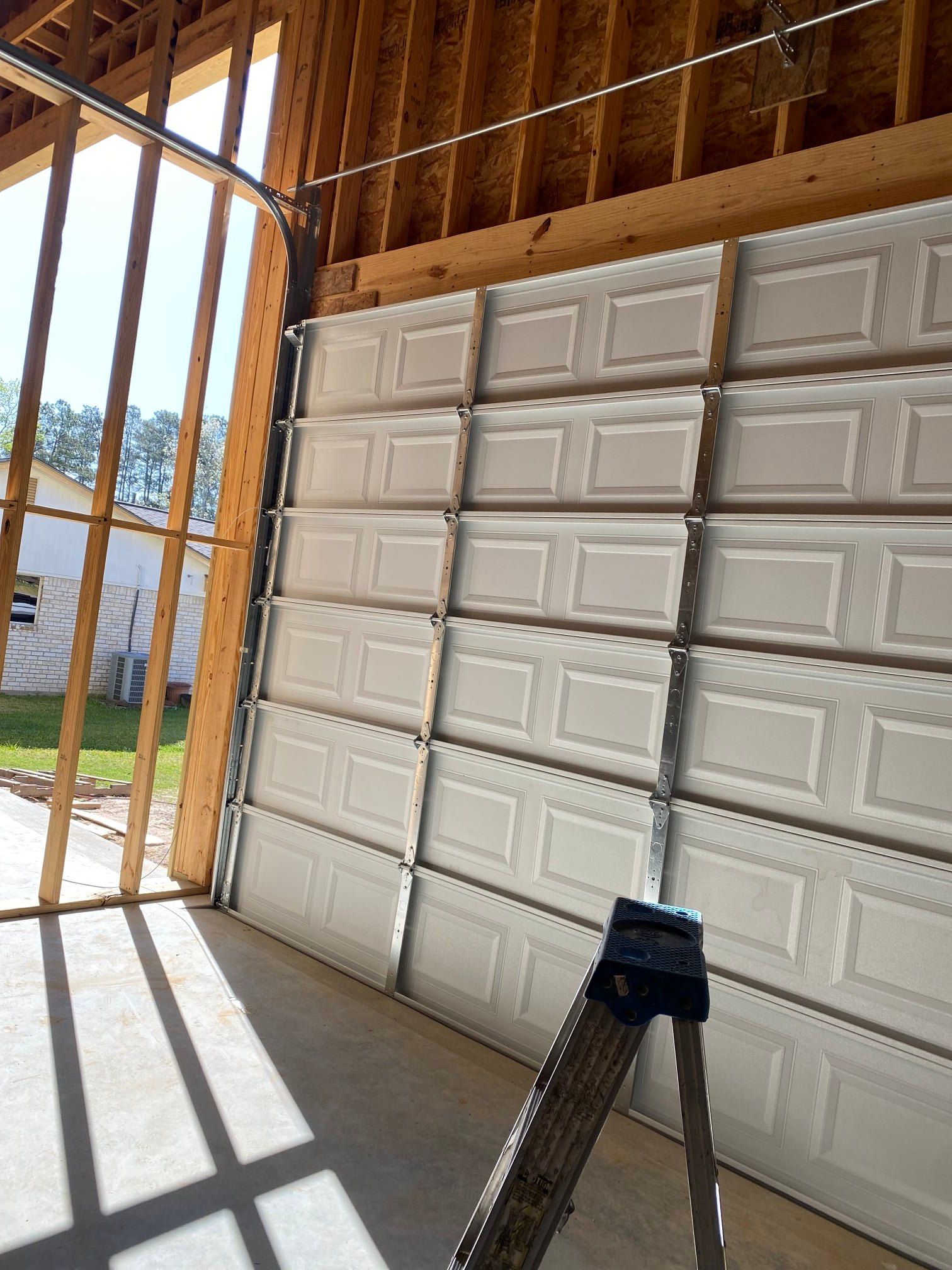 White multi-paneled garage door installed in a wooden garage frame with a ladder standing in the foreground.