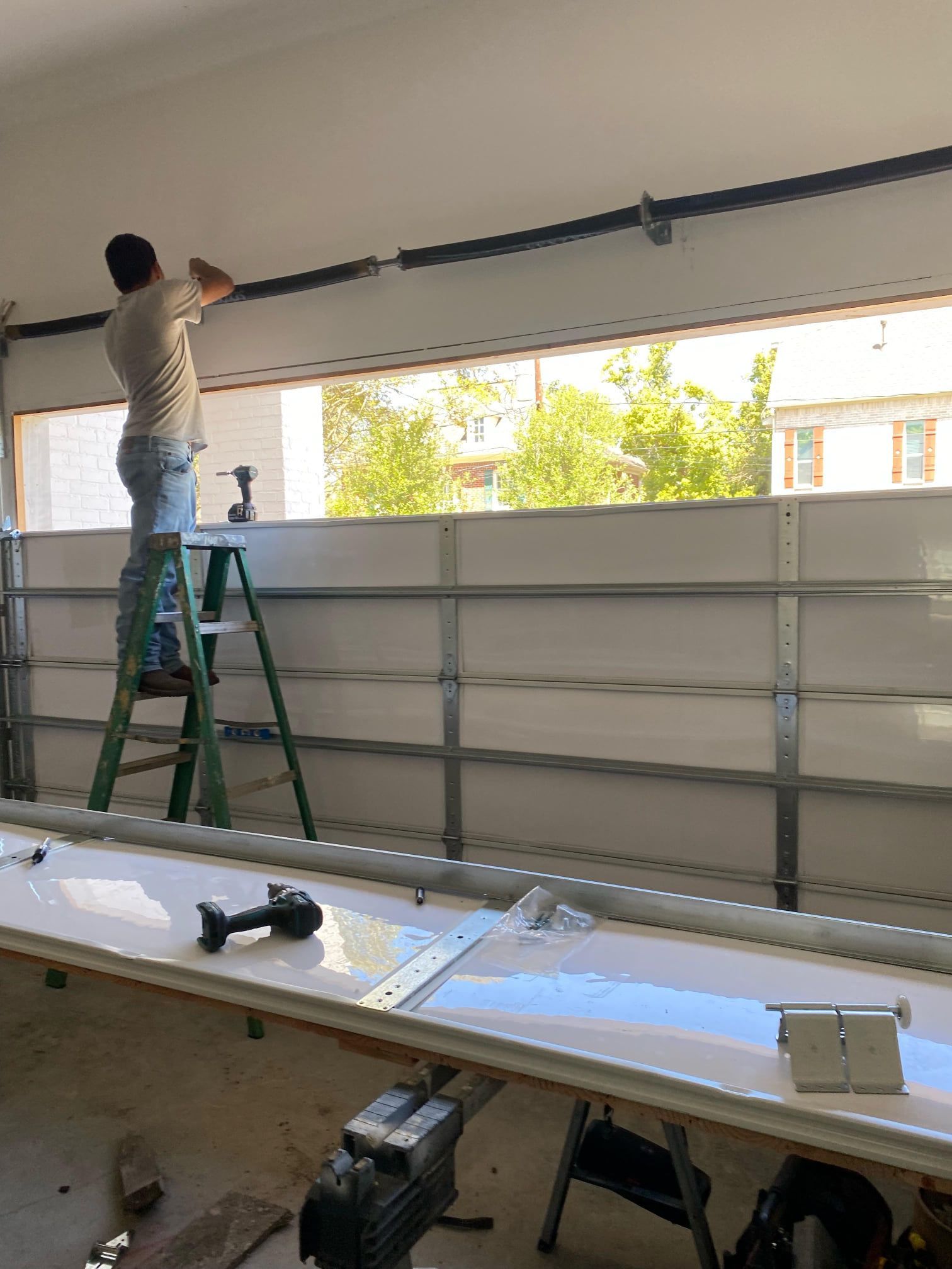 A person on a stepladder installs a garage door spring above an open garage door frame with white panels in the foreground.