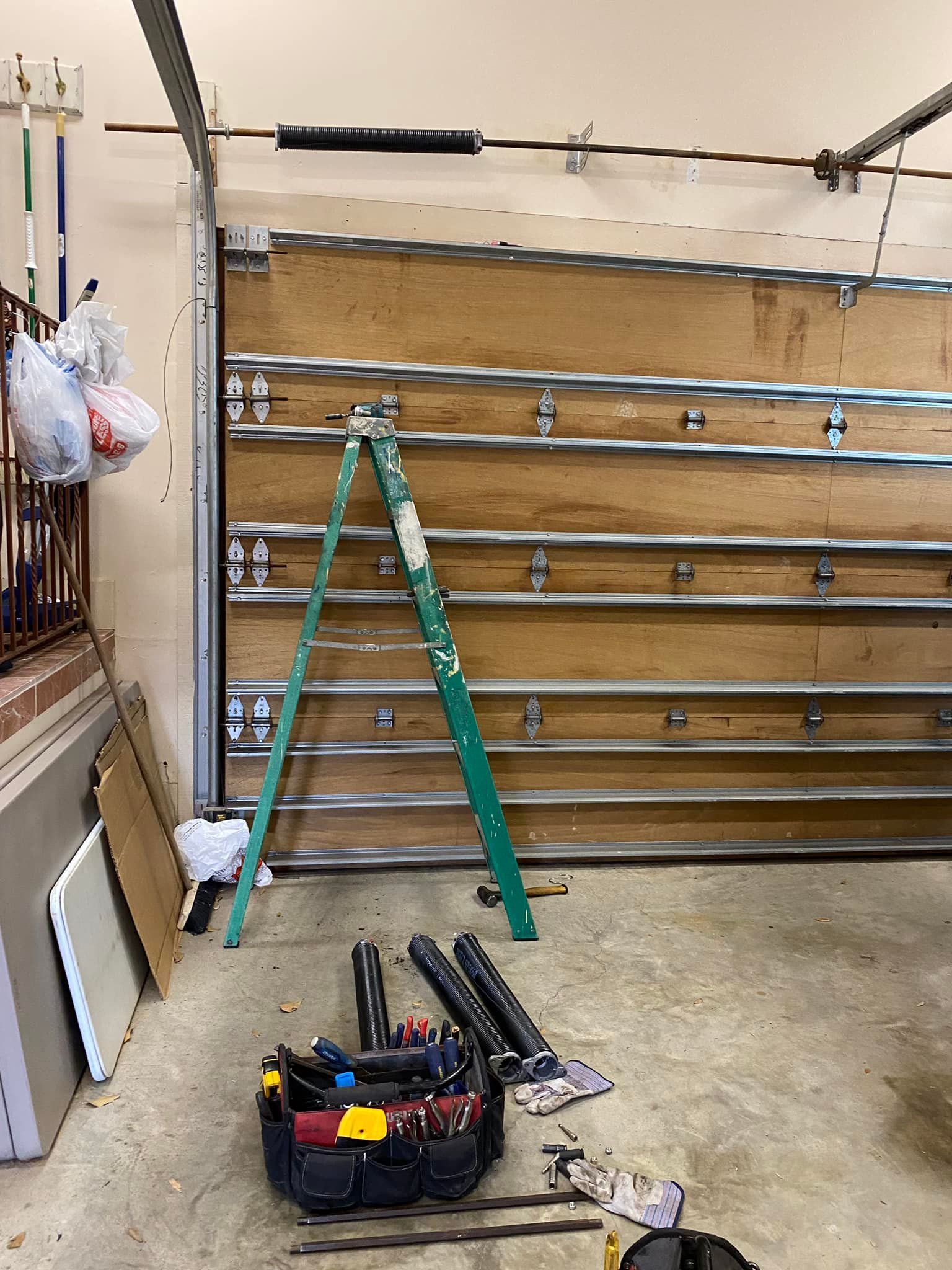 A garage door interior with a green step ladder standing in front of it and a tool bag resting on the floor.