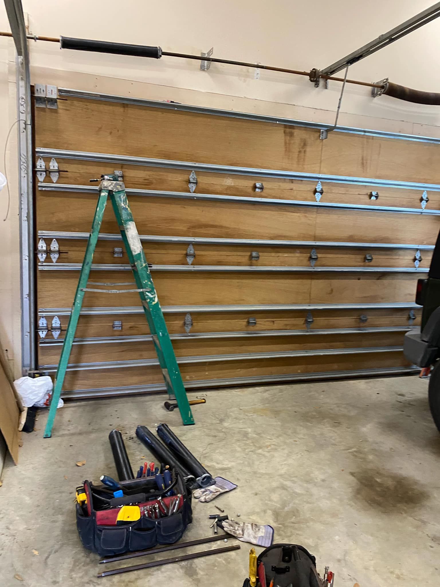 A ladder stands in front of a residential garage door with exposed metal support struts; tool bags sit on the floor nearby.