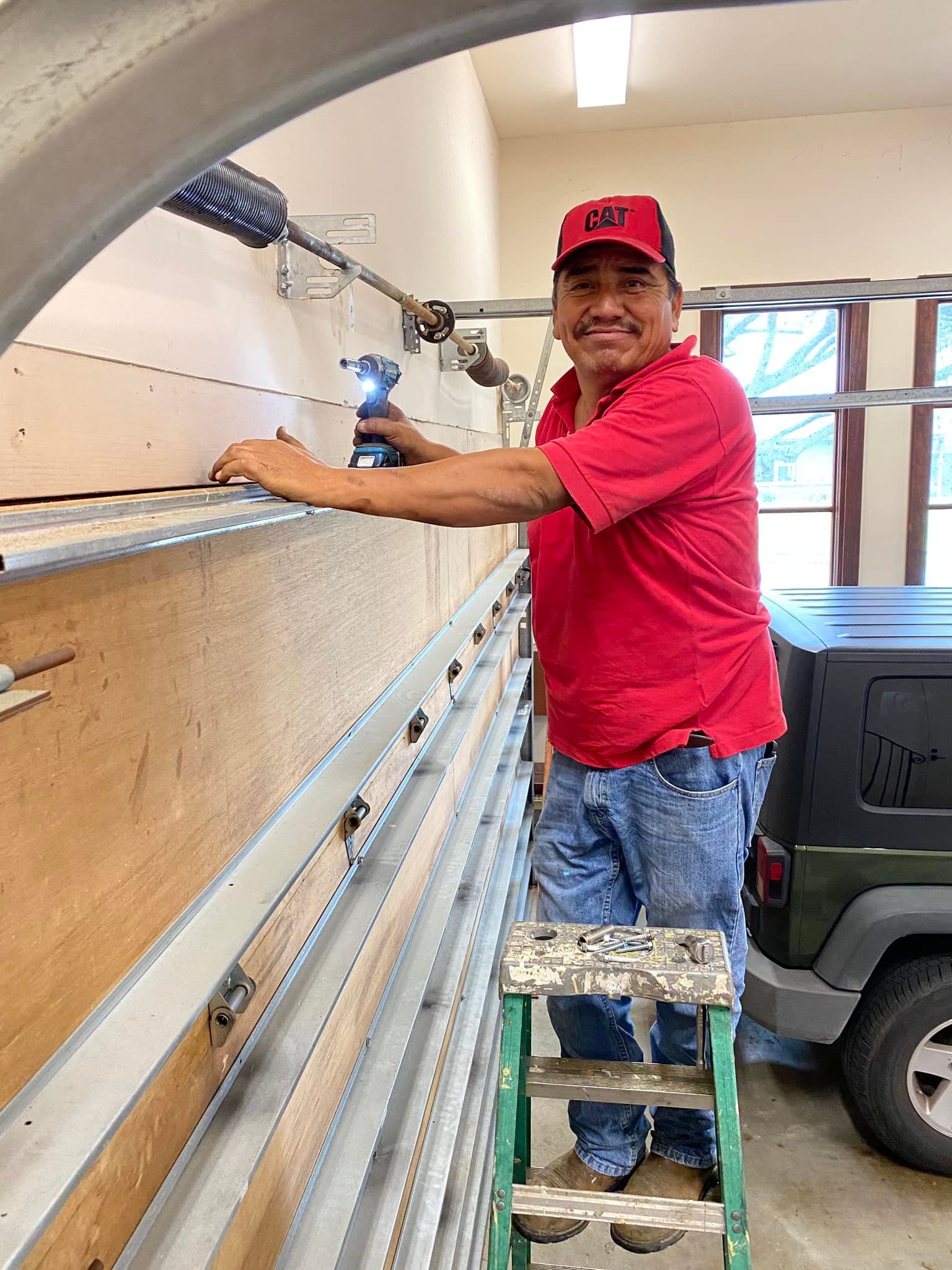A person wearing a red shirt and hat stands on a stepladder while using a power drill to install a garage door track.