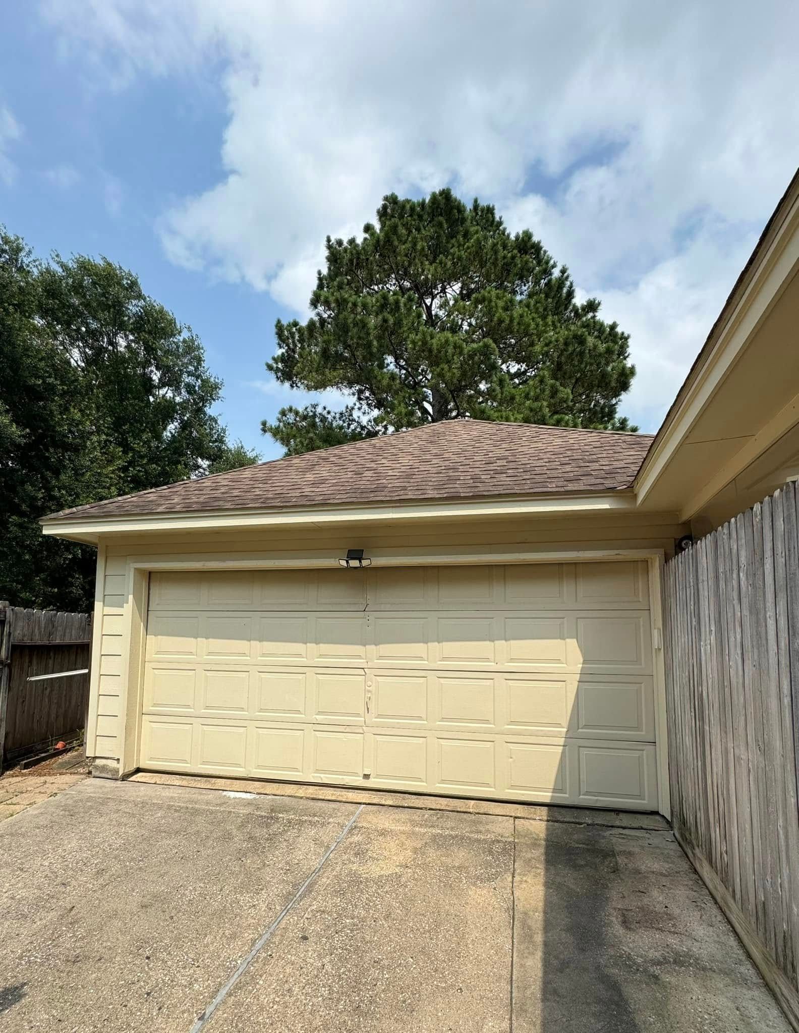 A beige garage door with horizontal dents in a driveway, set against a green tree and a blue sky with white clouds.