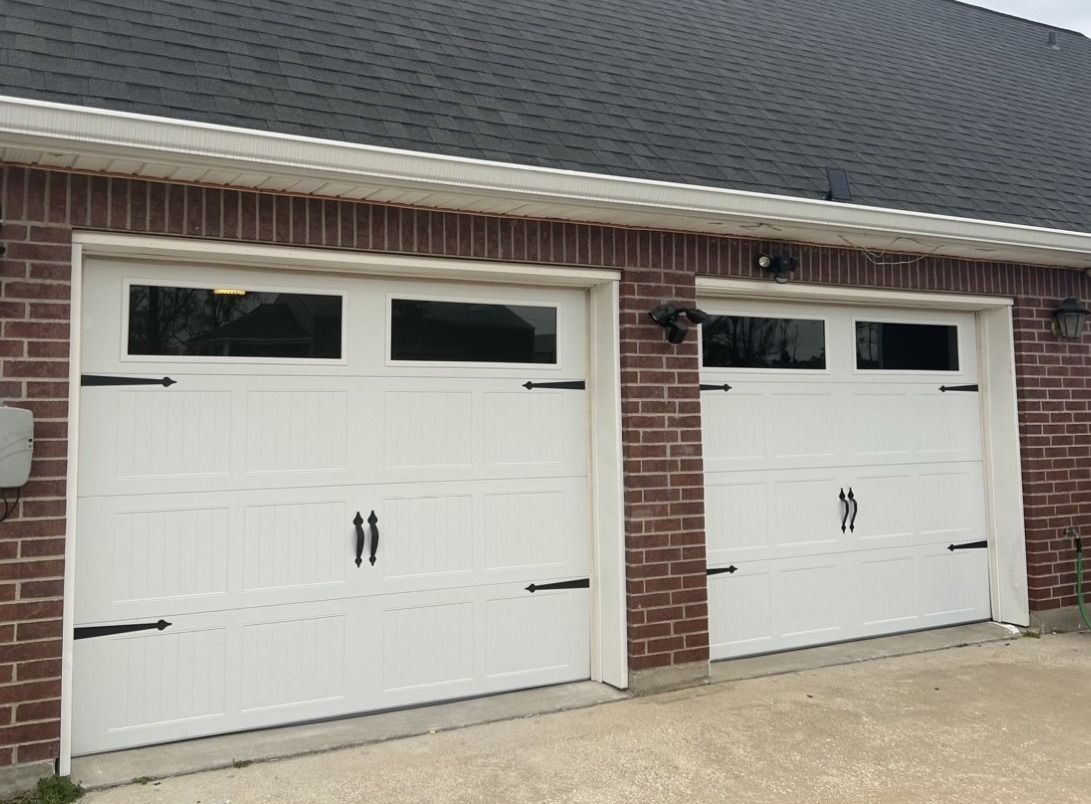 Two white residential garage doors with rectangular windows and black decorative handles, set in a red brick wall.
